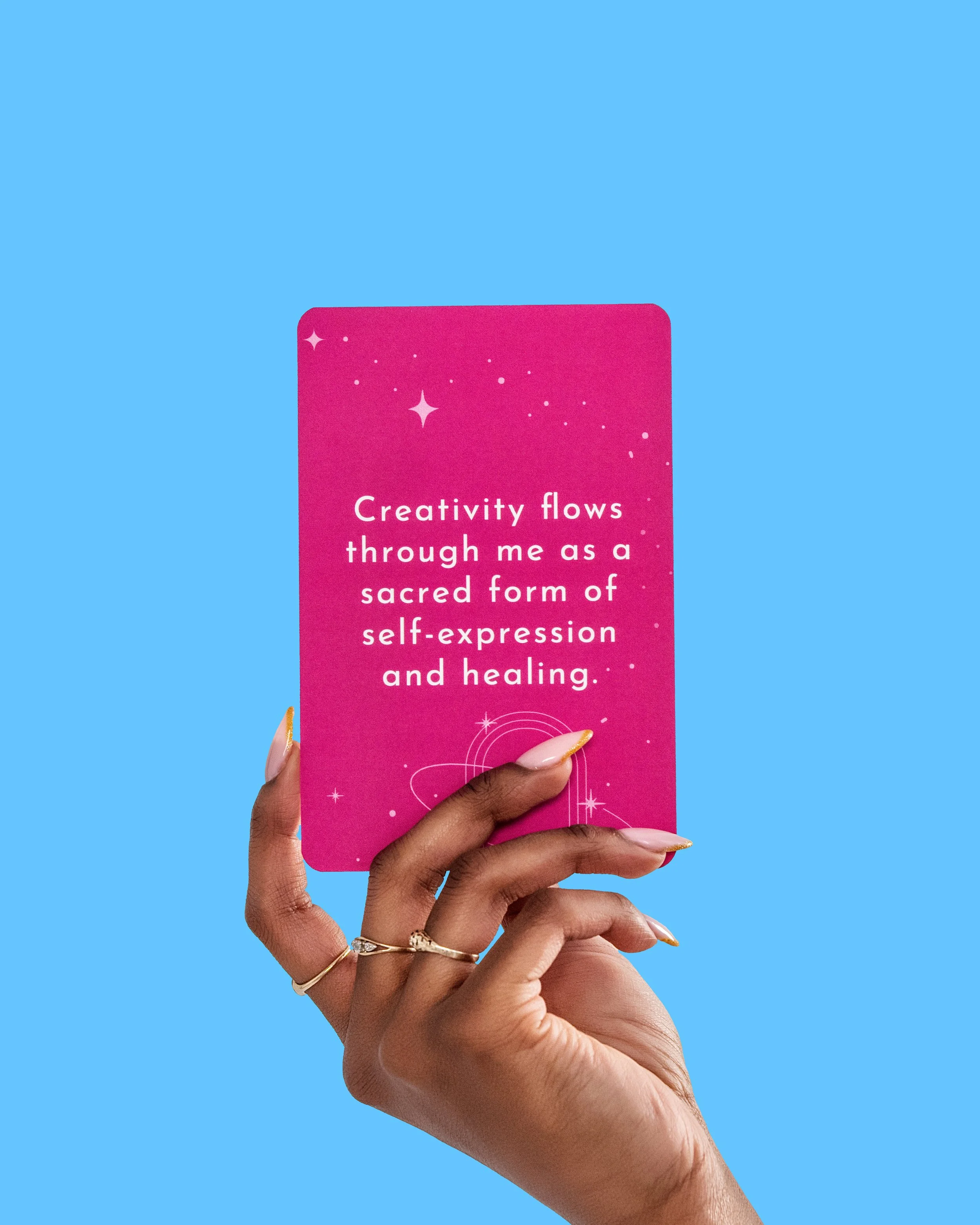 A hand with rings holding up a pink card with white text that reads, "Creativity flows through me as a sacred form of self-expression and healing" against a light blue background.