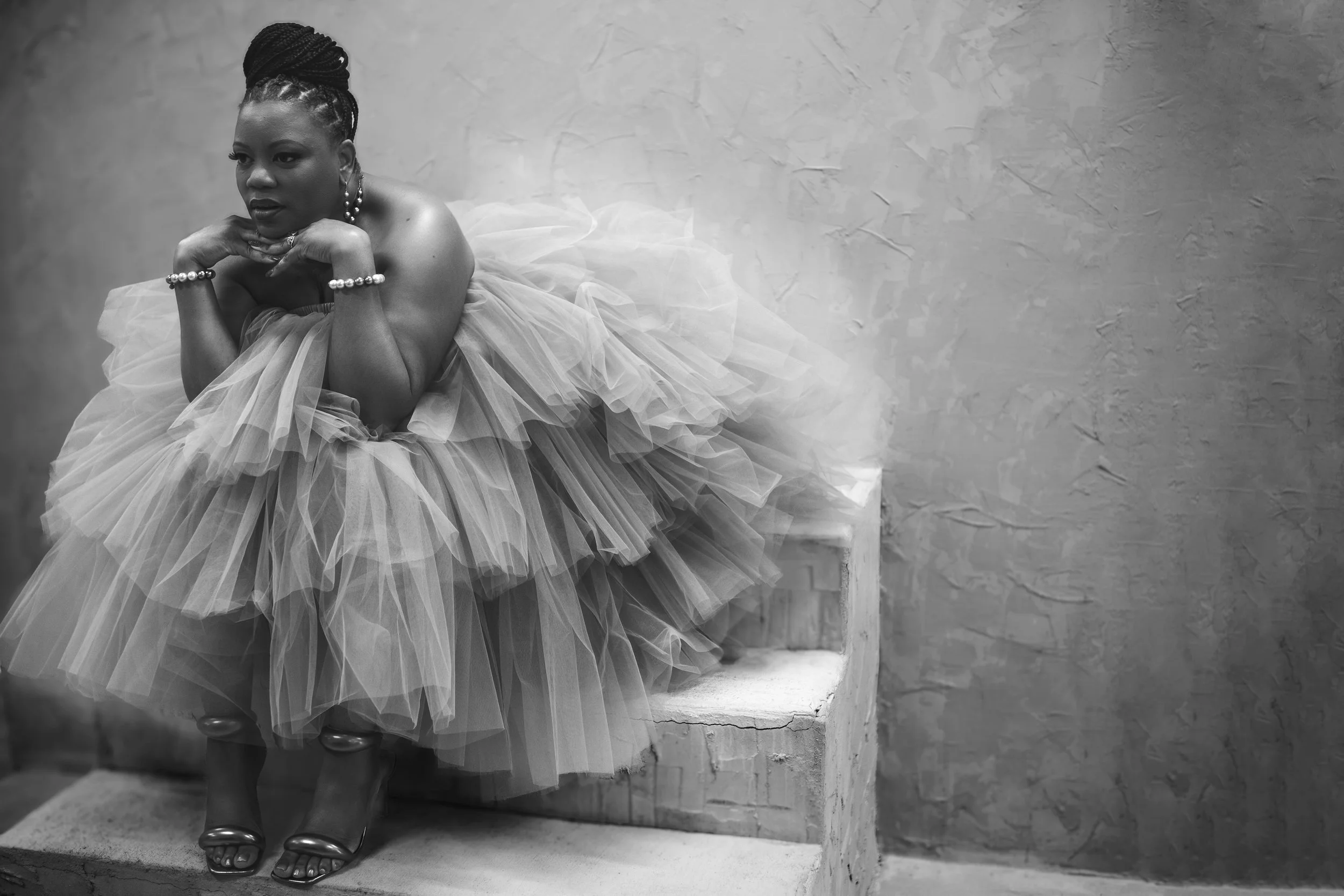 A woman dressed in a romantic, layered tulle gown sitting on stairs, with her hands resting under her chin, wearing jewelry, against a textured wall.