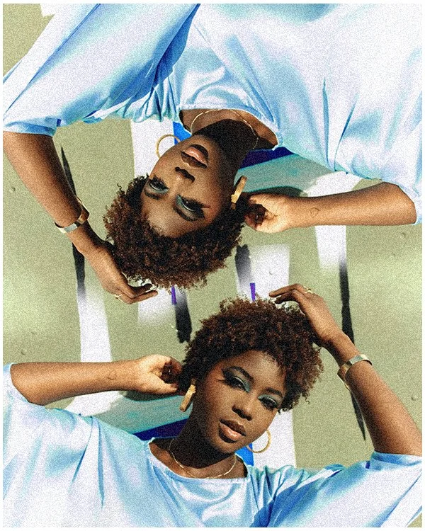A woman with curly hair and makeup, wearing a light blue jacket, is lying on her back on a reflective surface, with her arms bent and hands touching her hair, her eyes are closed or slightly open.
