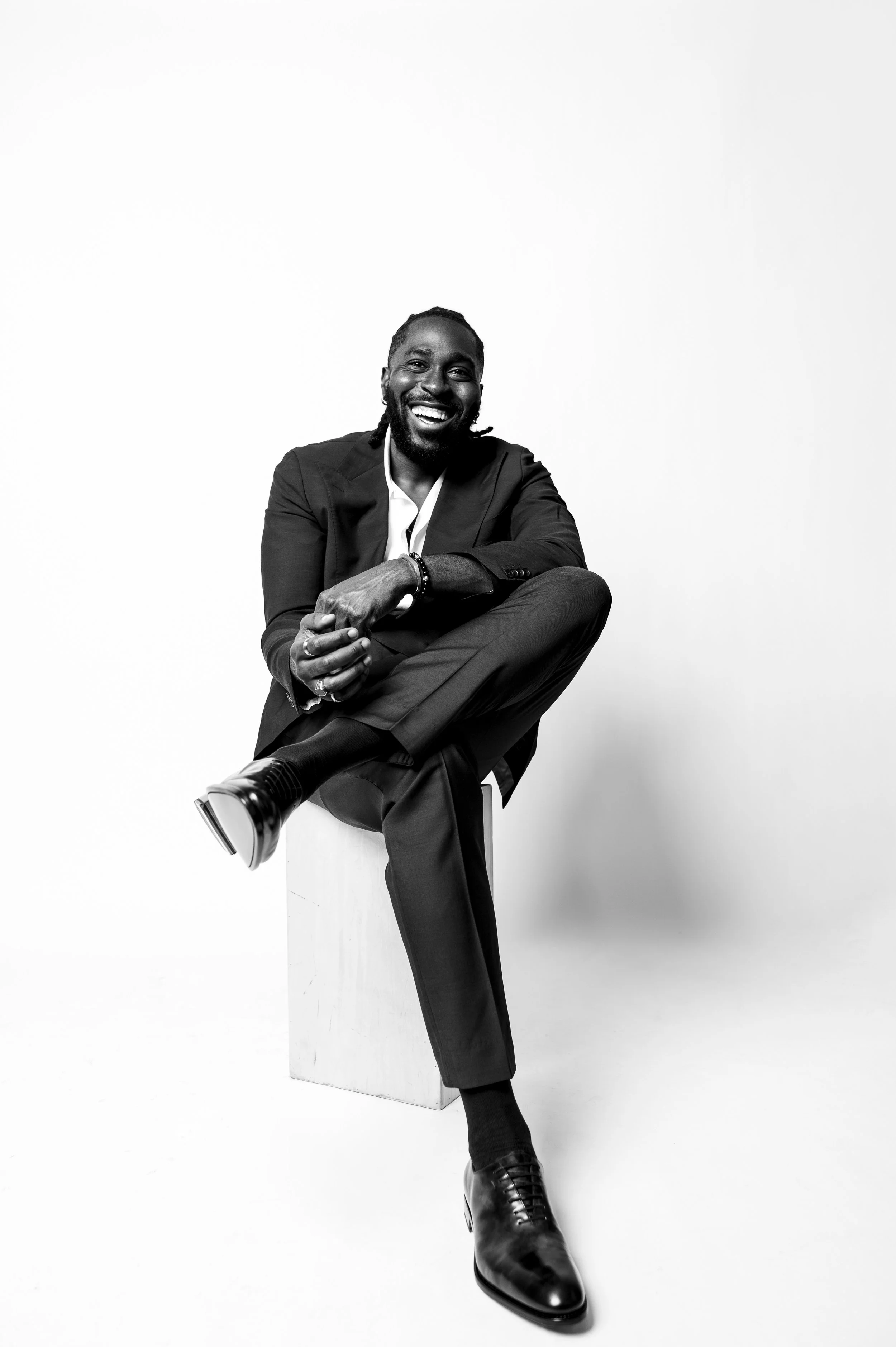 Black and white photo of a man in a suit sitting on a block, smiling, with one leg crossed over the other, against a plain white background.