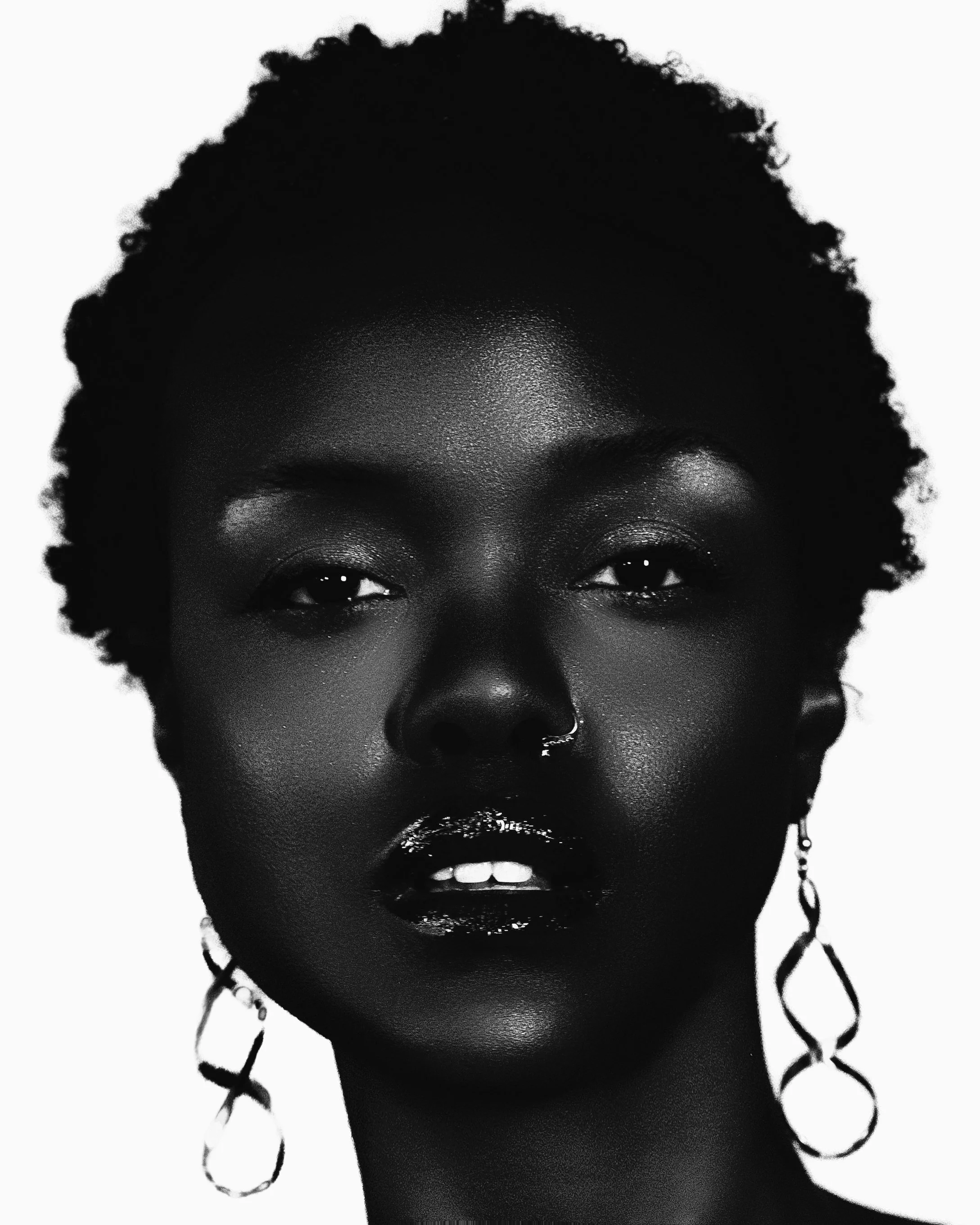 Black and white portrait of a woman with short, textured hair, wearing large hoop earrings, with makeup, and a nose piercing.