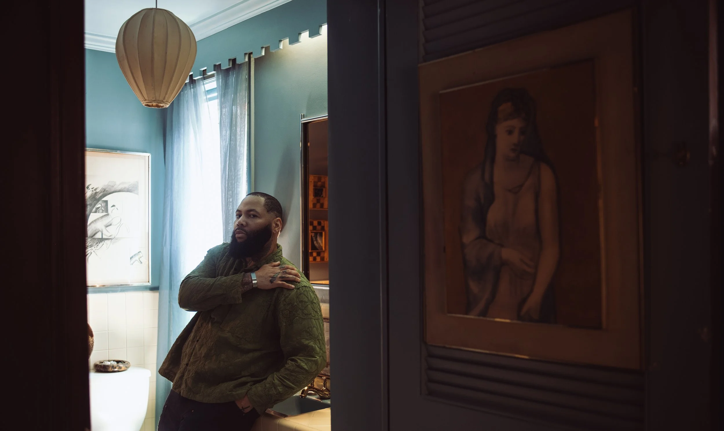 A man with a beard and tattoos leaning against a sink in a bathroom, with art and a window with curtains behind him