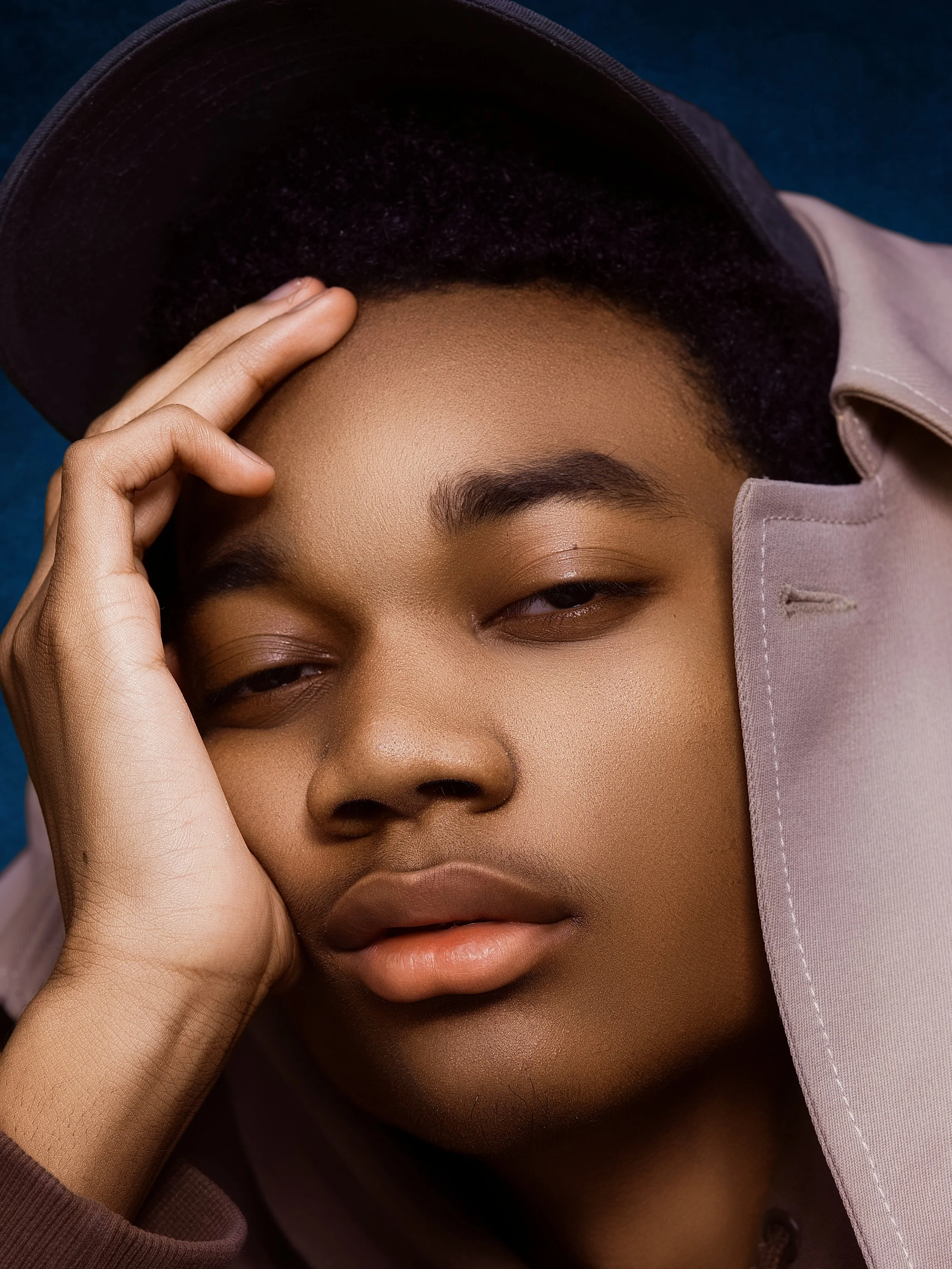 Close-up portrait of a young person with dark hair, wearing a black cap and a light-colored jacket, resting their head on their hand with a neutral expression.