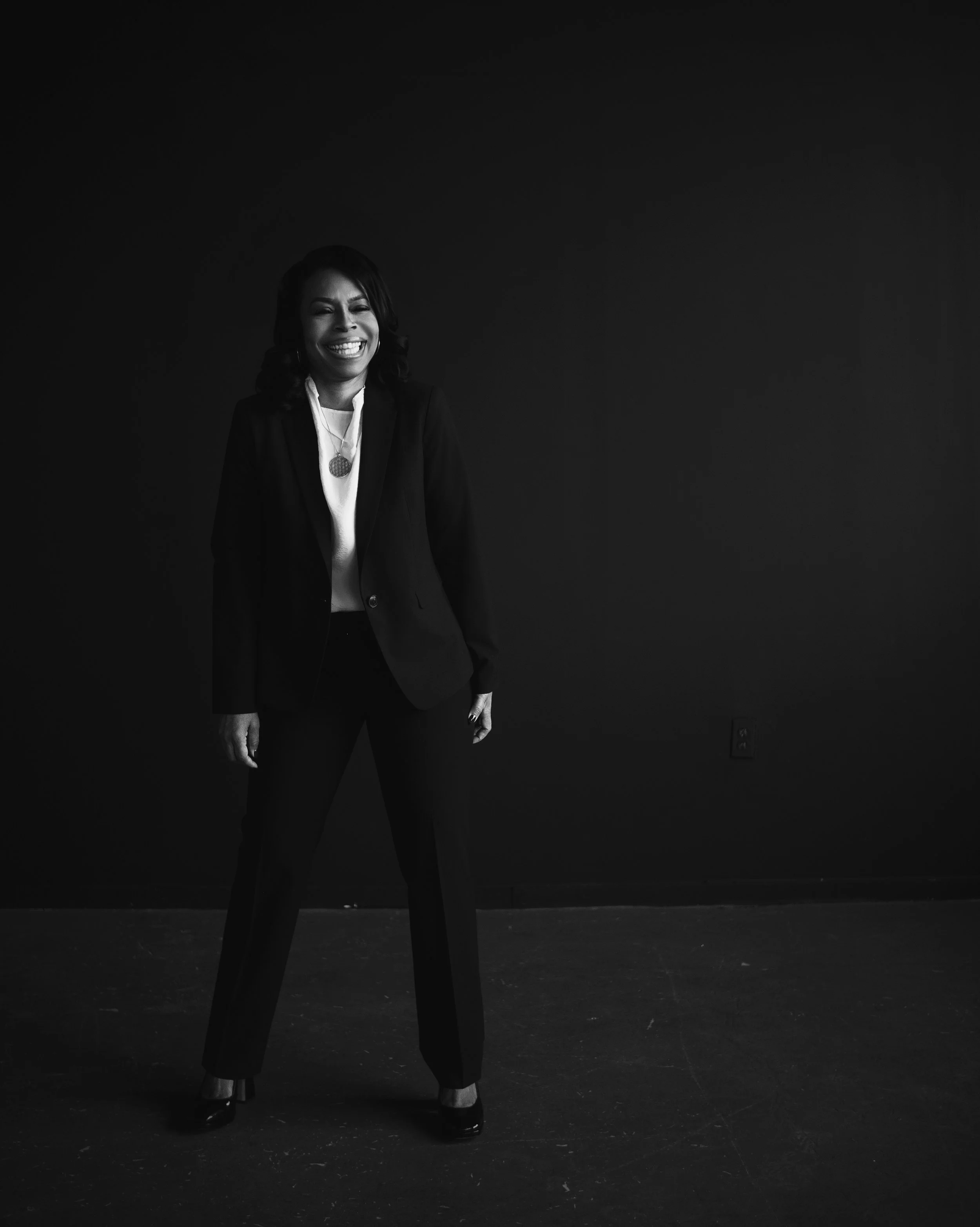 A woman in business attire smiling while standing against a dark background.
