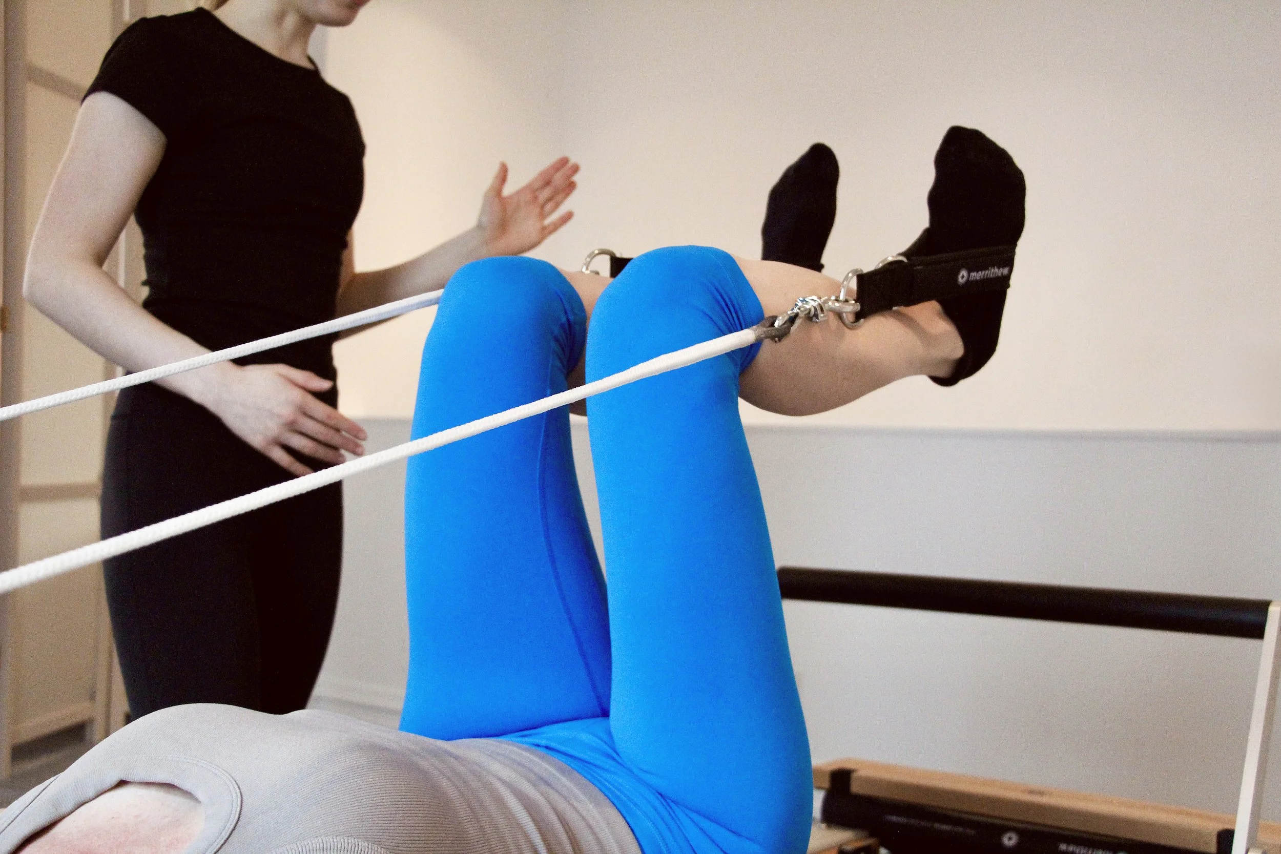 private client doing feet in straps on reformer with instructor spotting