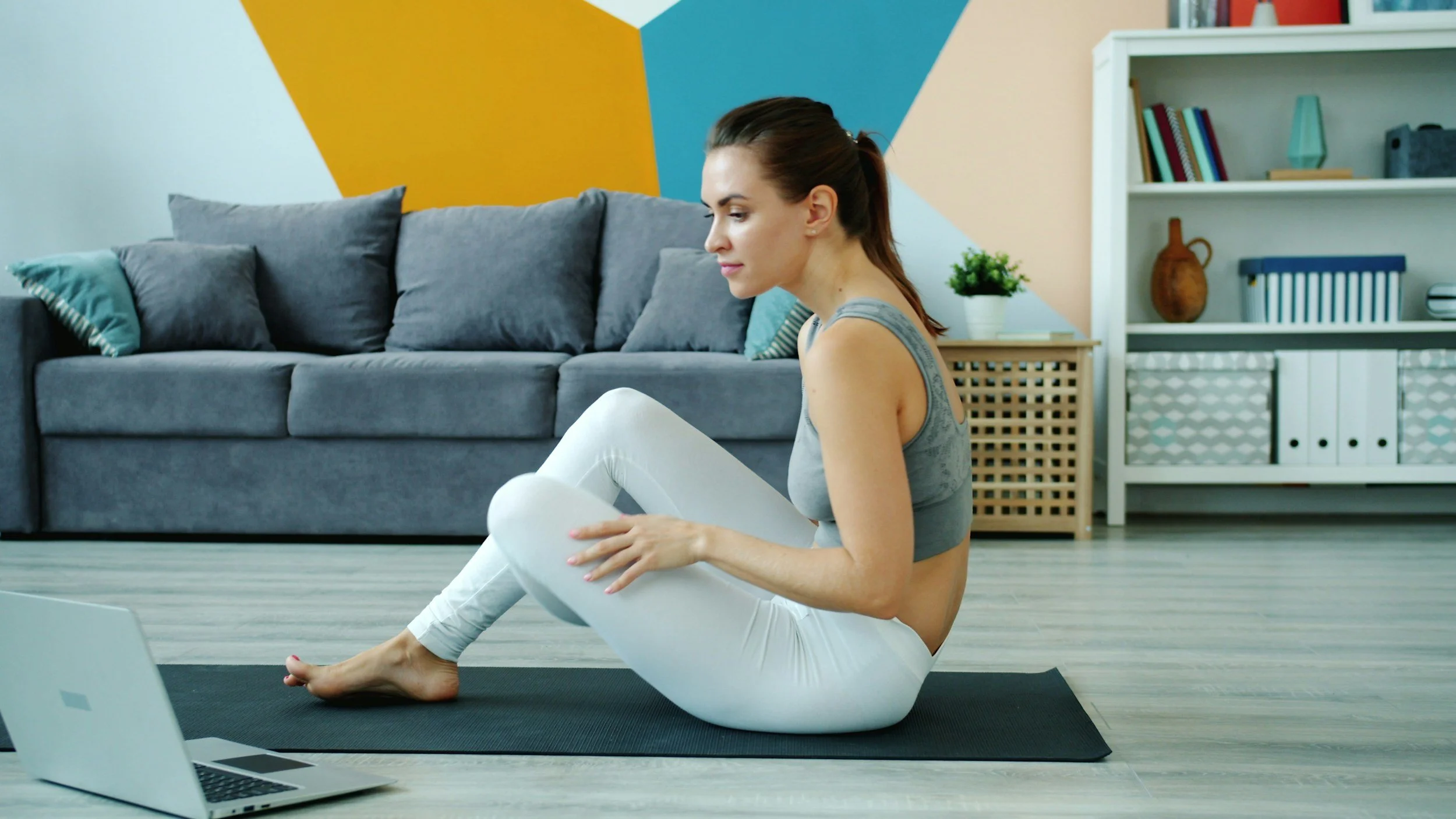 A woman in athletic wear practicing yoga or stretching on a mat in a living room with colorful wall art, a gray sofa, and a white bookshelf.