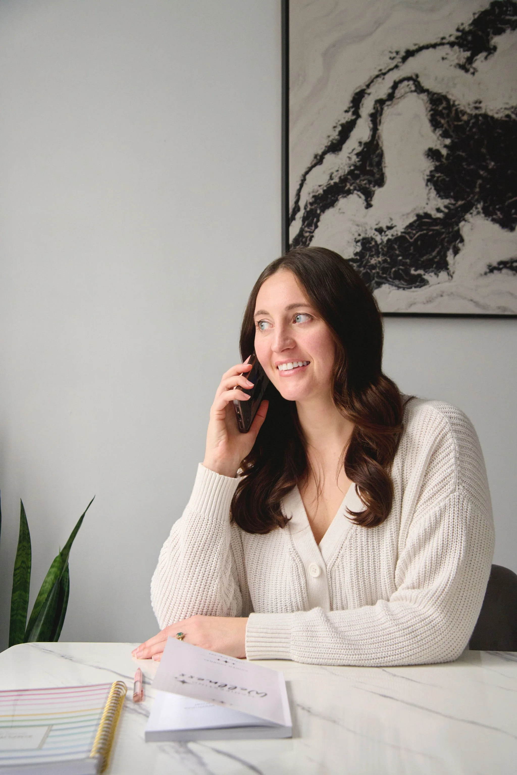 A woman with long, dark, wavy hair smiling while talking on her cell phone, sitting at a white marble table with colorful notebooks, papers, a pink pen, and a plant nearby, against a plain white wall with a black and white abstract painting.