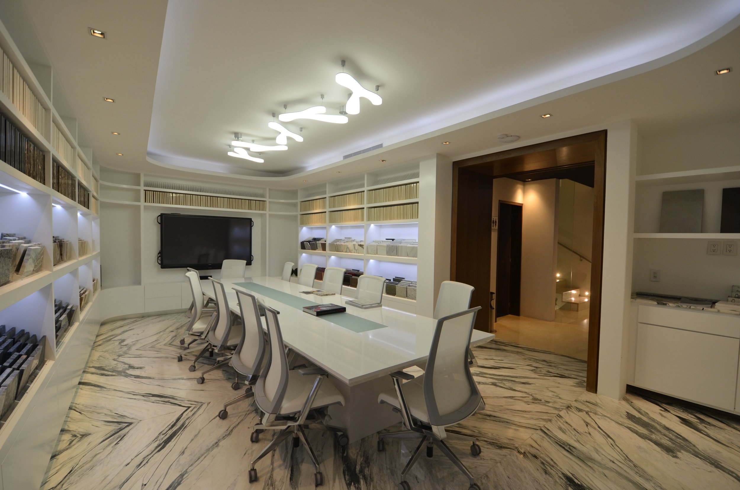 Modern conference room with white walls, a long white table with nine white chairs, built-in bookshelves with books and files, a large flat-screen TV, marble flooring, and a ceiling light fixture with abstract shapes.