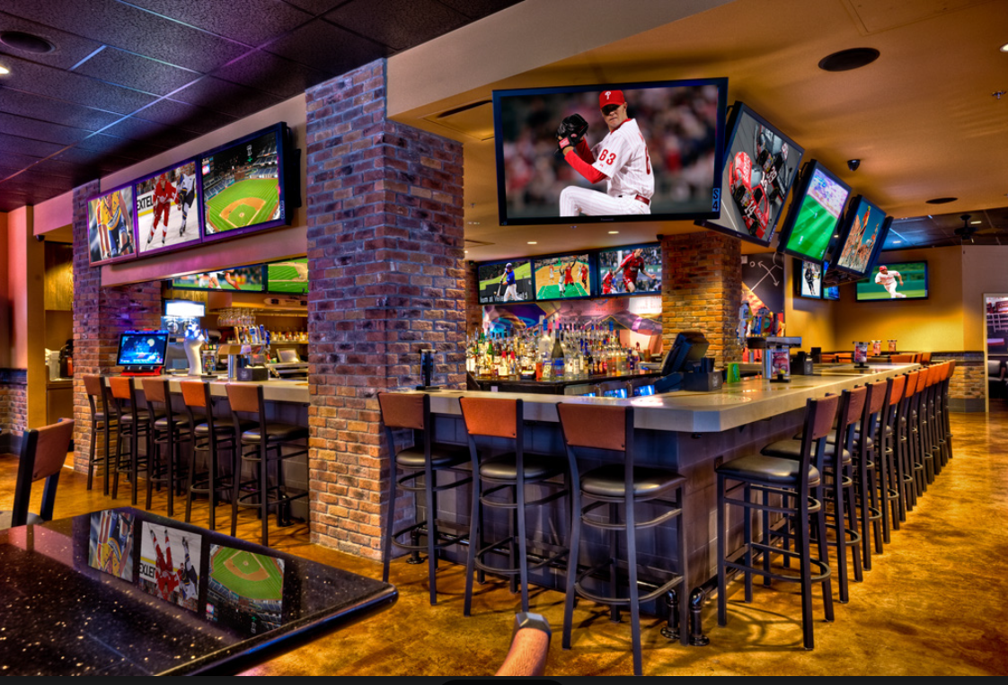 Sports bar with multiple flat-screen TVs showing various sports, a long bar counter with chairs, and brick pillars.