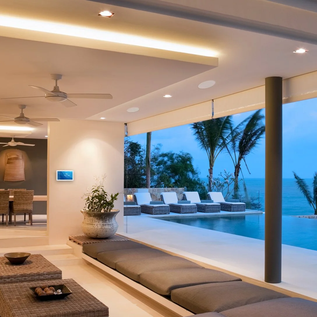 Indoor lounge area with ceiling fans and ambient lighting, overlooking an outdoor pool and patio with lounge chairs and palm trees, overlooking the ocean at dusk.