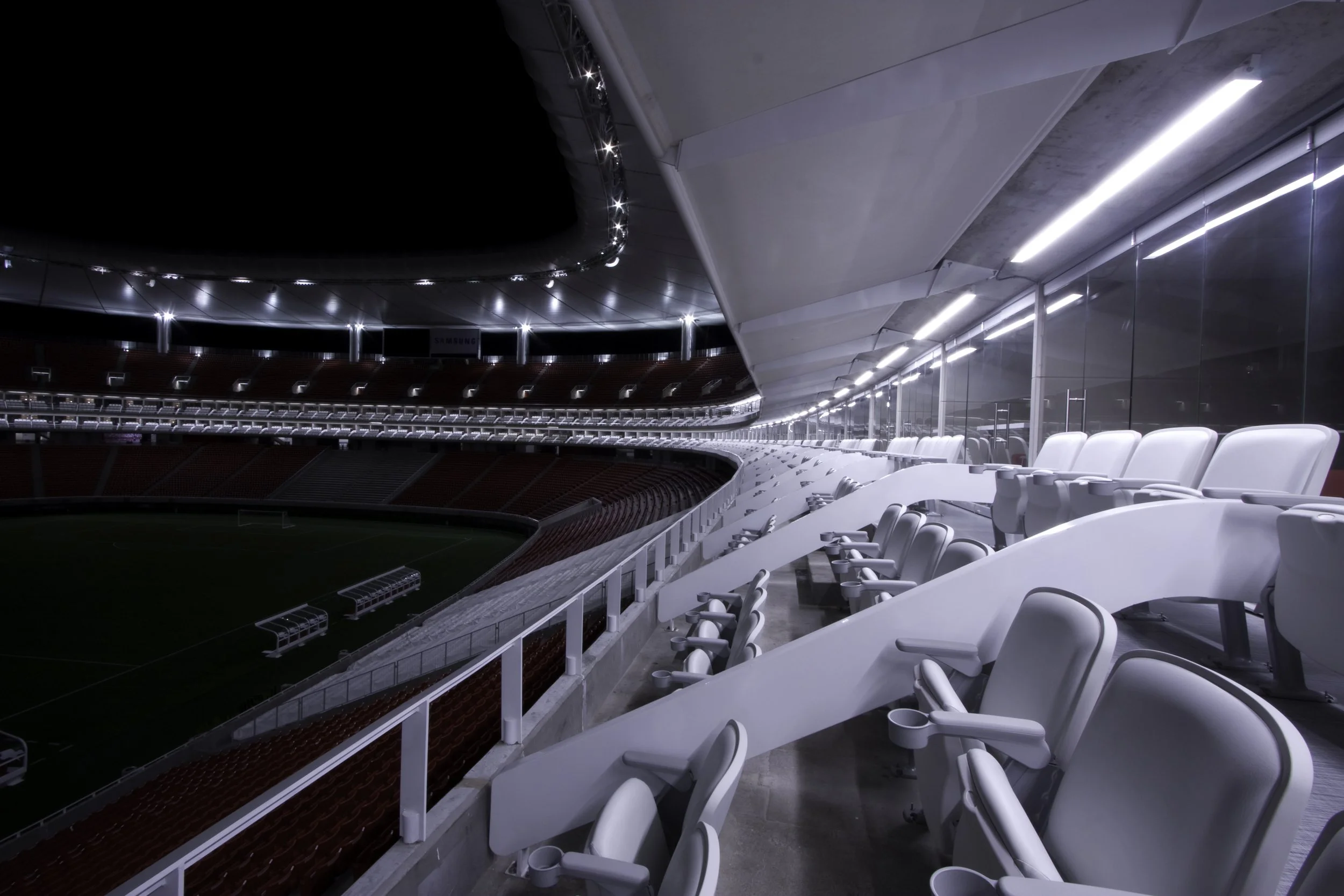 Empty stadium seats and a soccer field in the background, with a modern enclosed balcony area with white chairs along large glass windows.