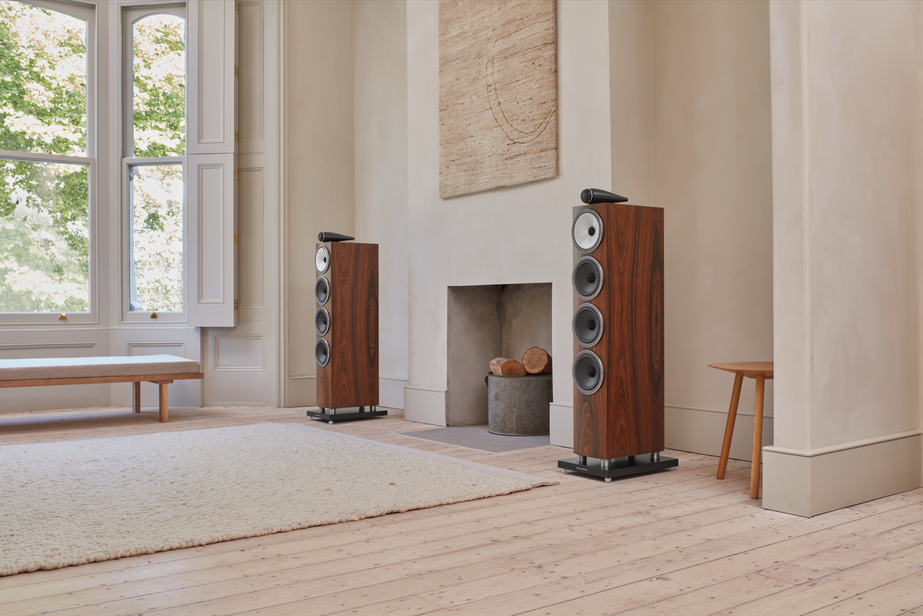 Living room with large window, beige walls, fireplace with logs, two tall wooden speakers, small wooden stool, and beige carpet.