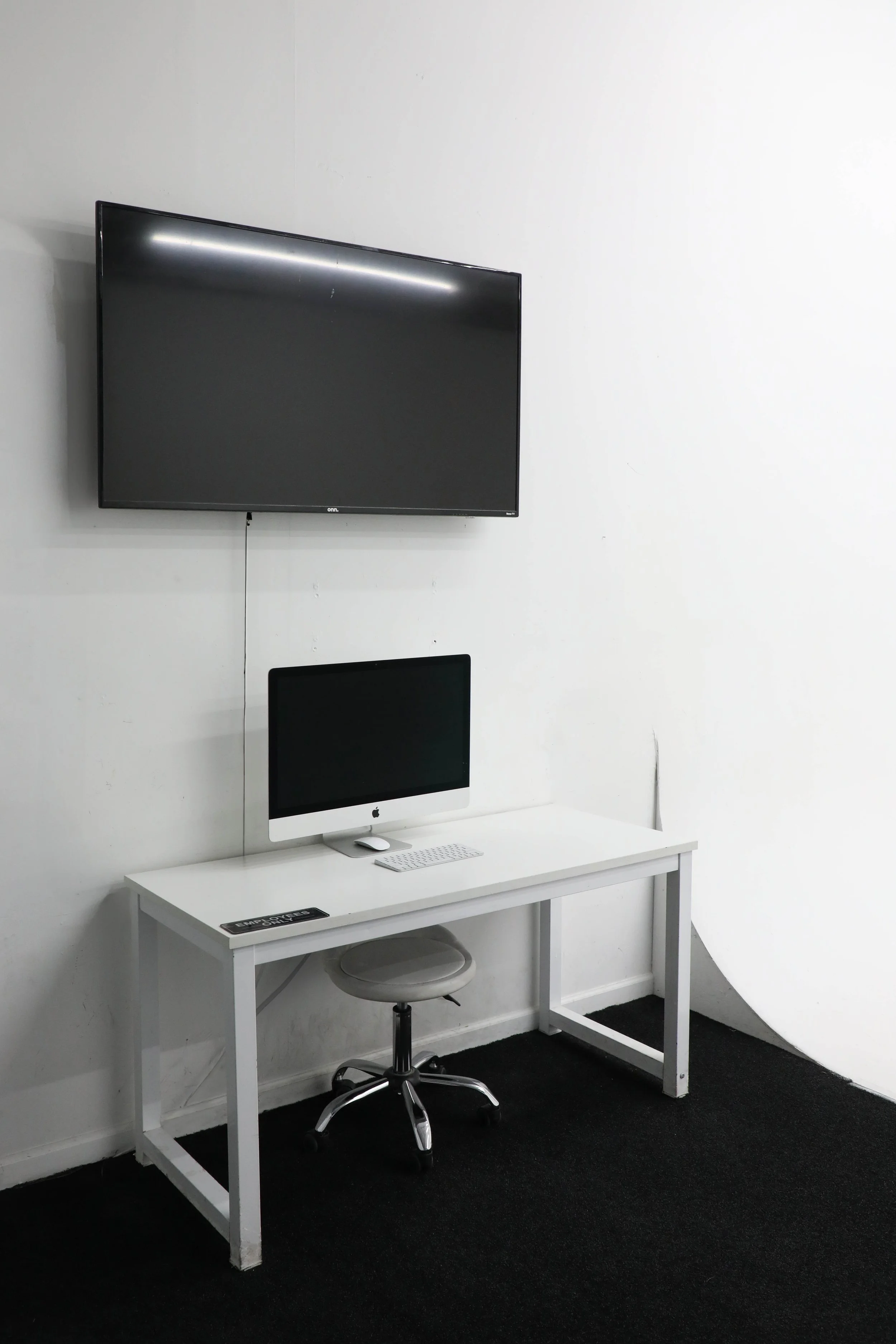 A modern workspace with a white desk, an iMac computer, a wireless keyboard, and a mouse. A wall-mounted flat-screen TV is above the desk, and a swivel chair is in front of the desk. The scene has a minimalist design with a black carpet and white wal