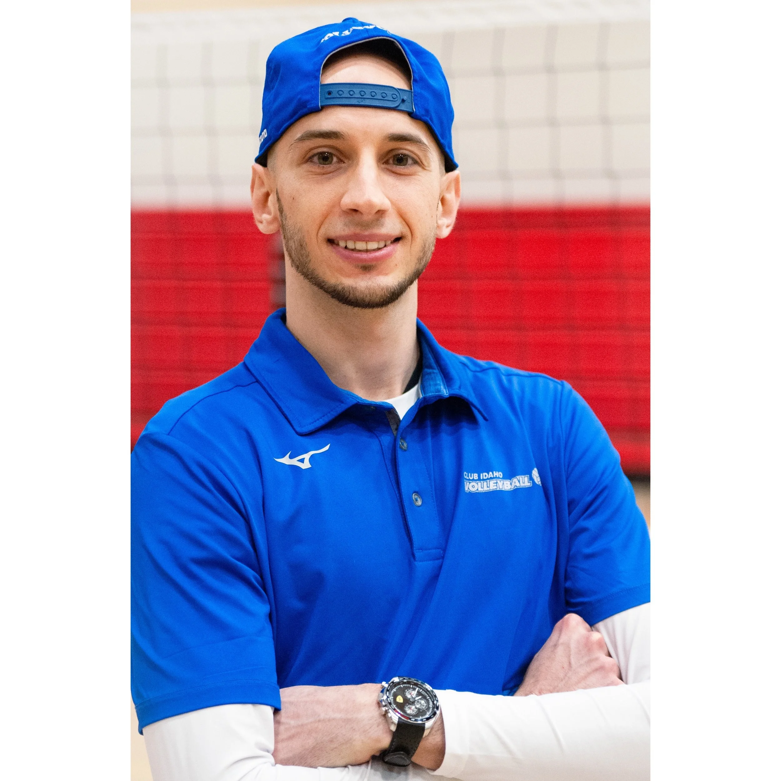 A young man in a blue volleyball uniform and cap, smiling with arms crossed, standing in an indoor gym with red and beige bleachers in the background.