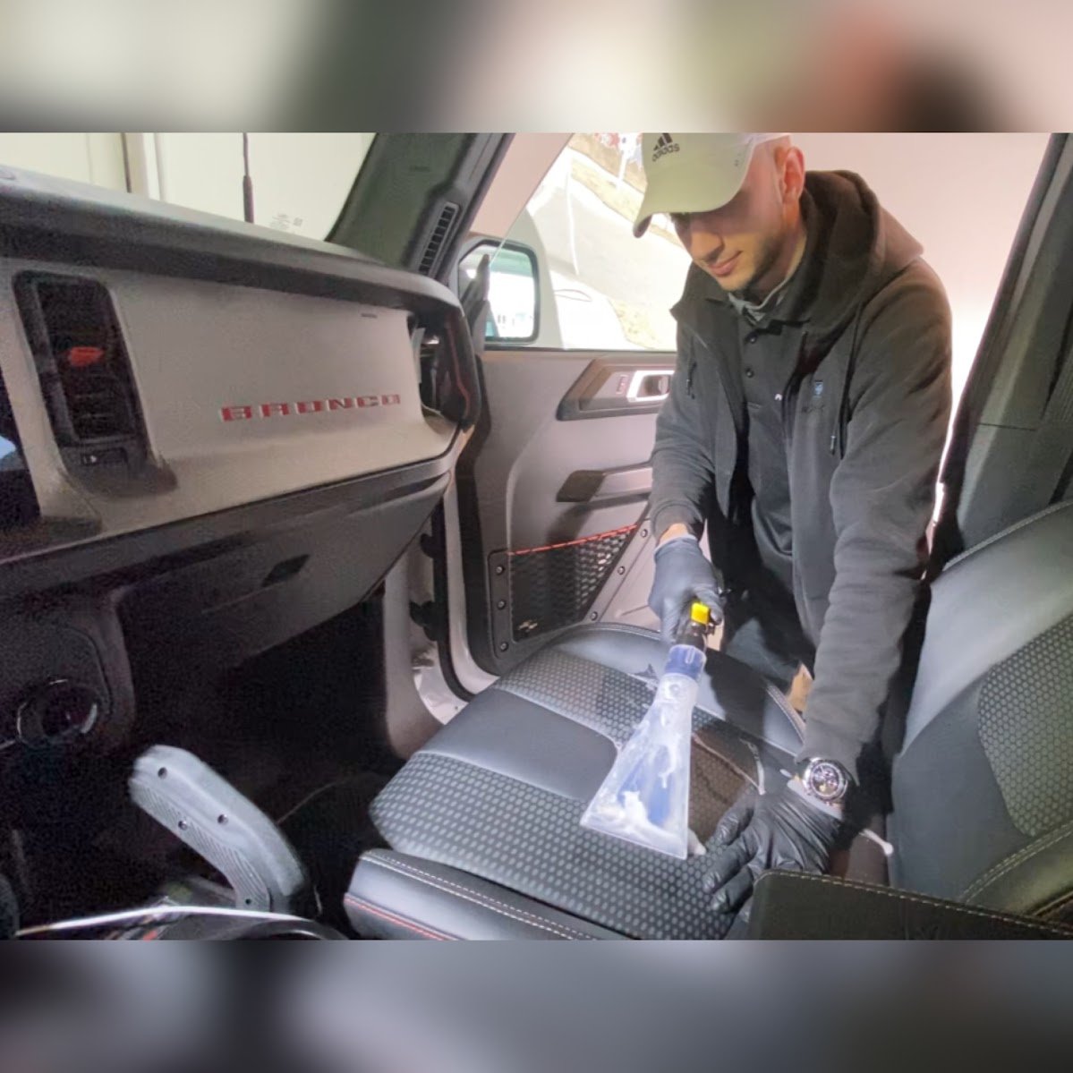 A man wearing a beige cap, glasses, gloves, and a dark jacket is spraying disinfectant on the seat inside a vehicle, likely a truck, with a spray bottle.