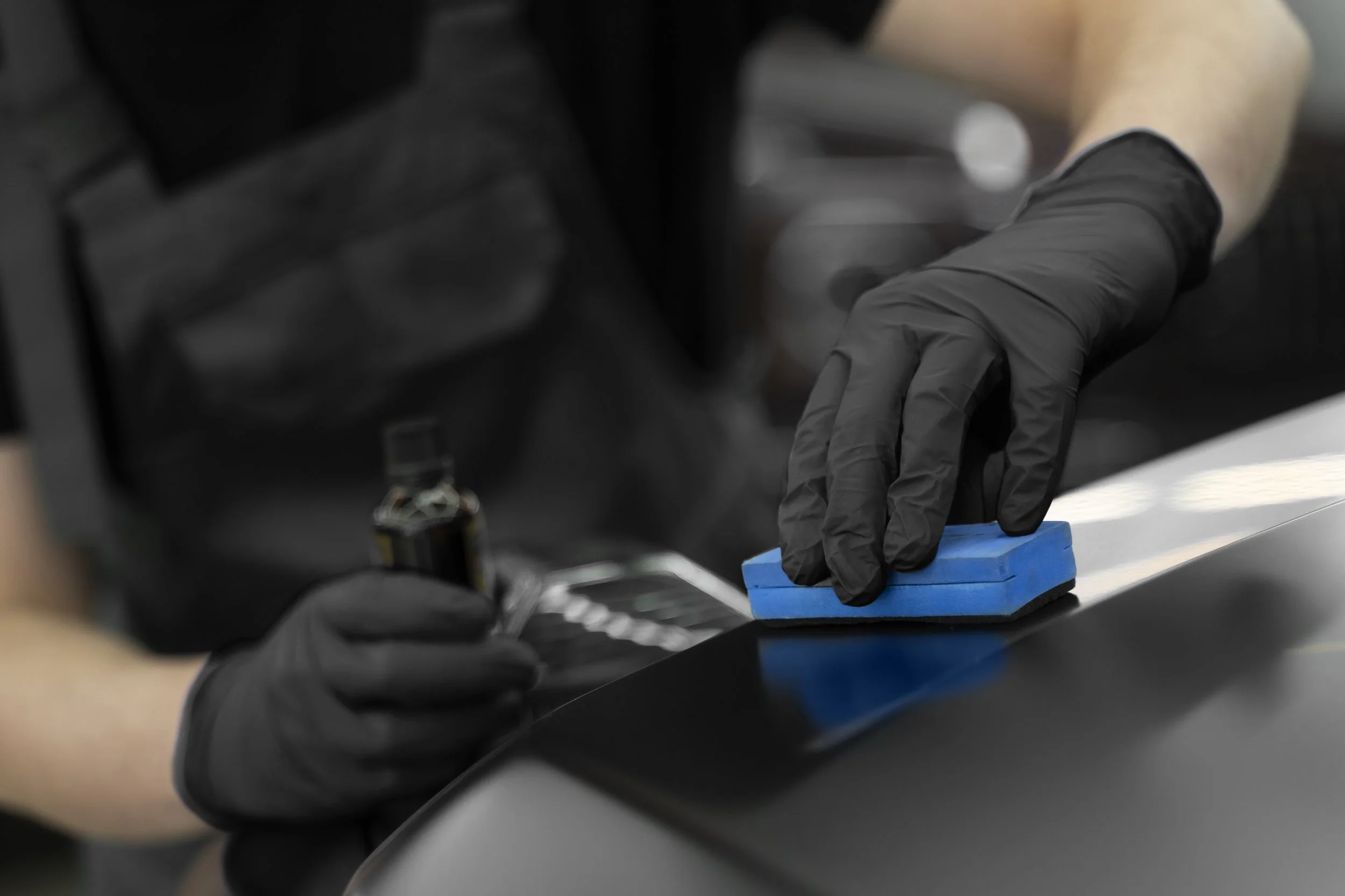 Person wearing black gloves cleaning a car with a sponge and a spray bottle.