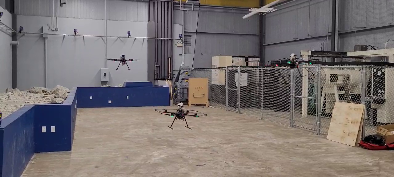 Two drones flying in an industrial warehouse with equipment and fencing.