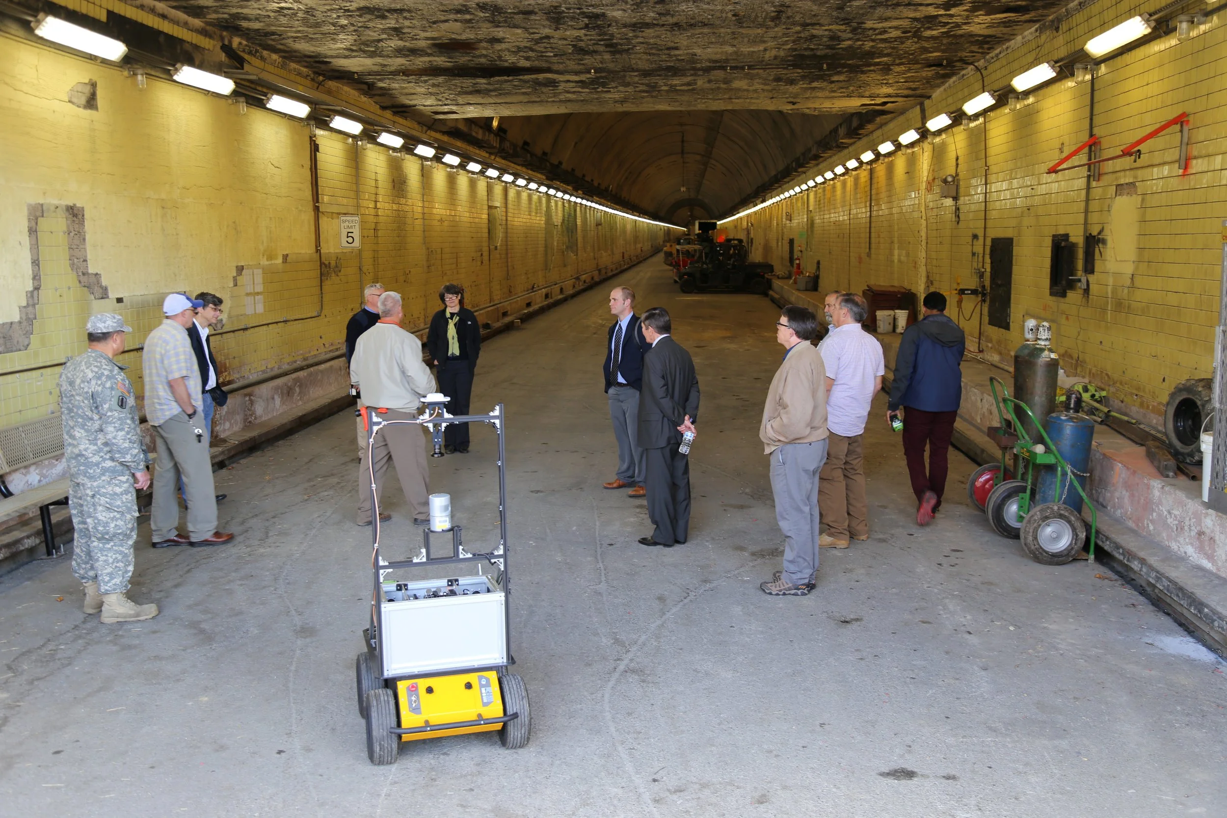 Group of people standing inside a tunnel with yellow tiles, some equipment on the floor, and a vehicle in the distance.