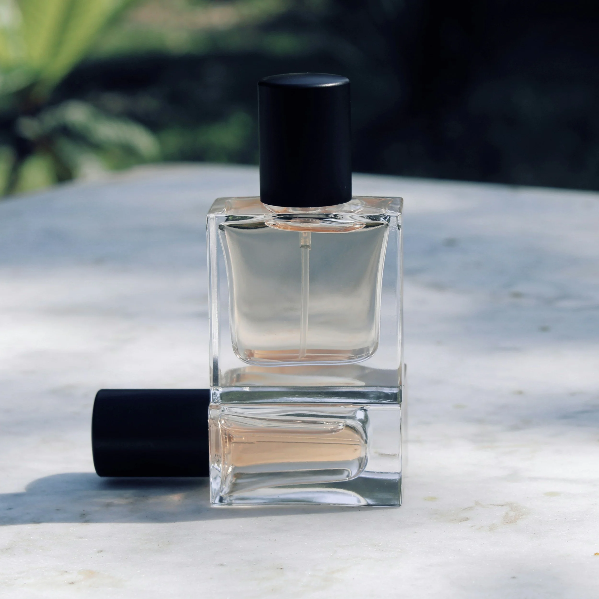 A clear glass perfume bottle with a black cap, placed on a white marble surface with a blurred outdoor background.
