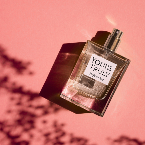 A perfume bottle labeled 'YOURS TRULY Perfume Bar' on a pink background with shadows and a black cap.