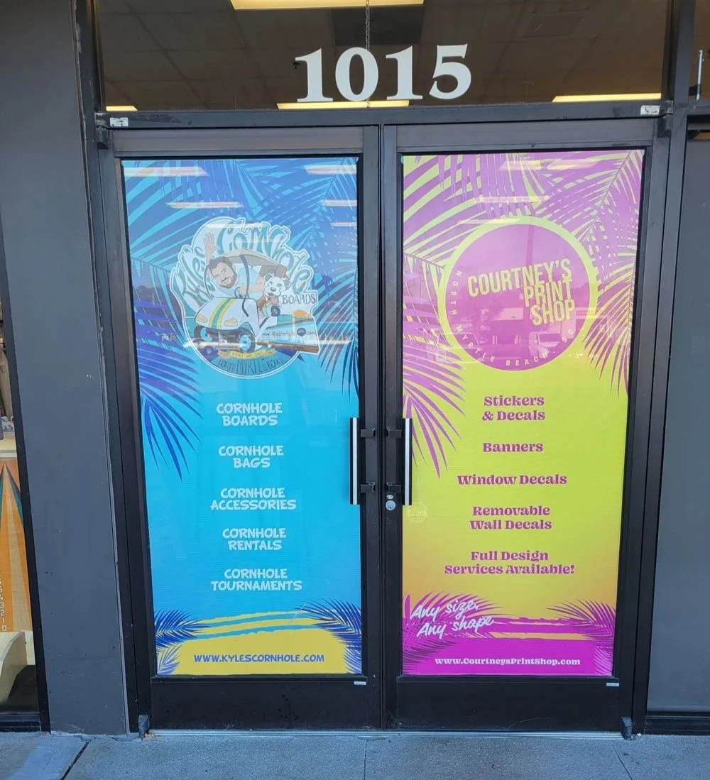 Storefront window decals for Kyle’s Cornhole Boards and Courtney’s Print Shop in North Myrtle Beach featuring colorful tropical palm designs, advertising cornhole boards