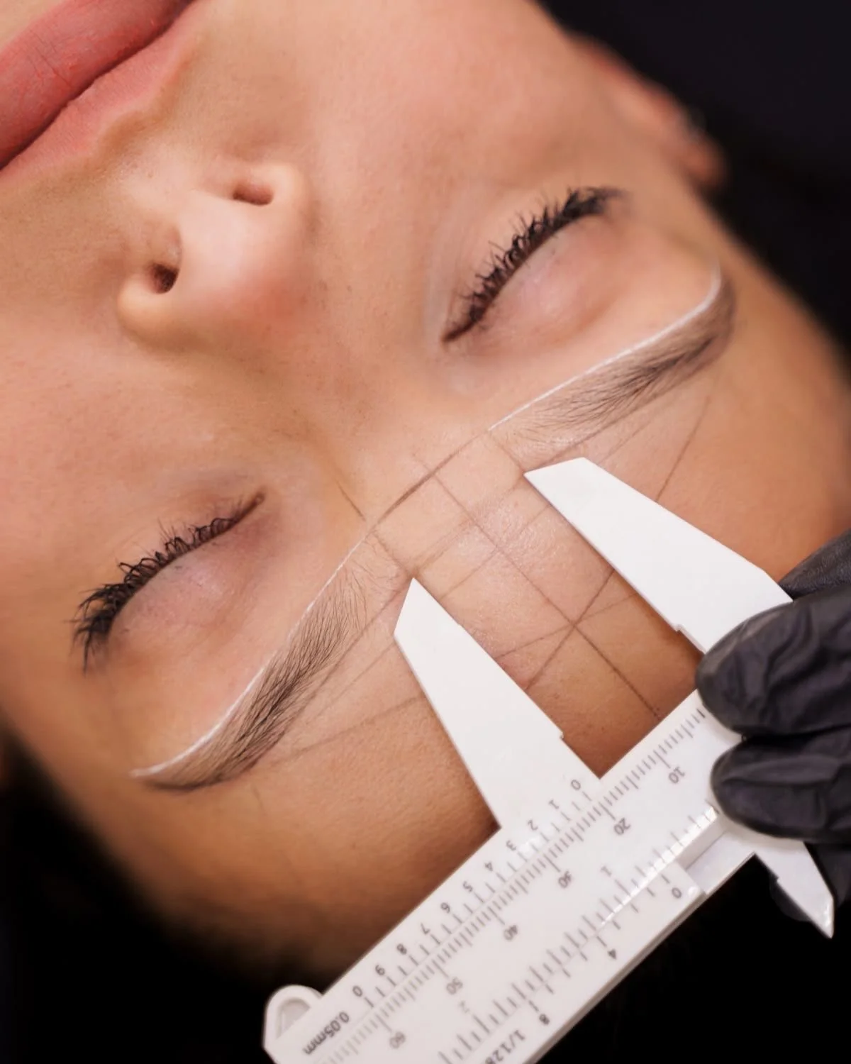 Close-up of a woman receiving a cosmetic tattooing or microblading procedure on her eyebrows, with a visible ruler guiding the process.