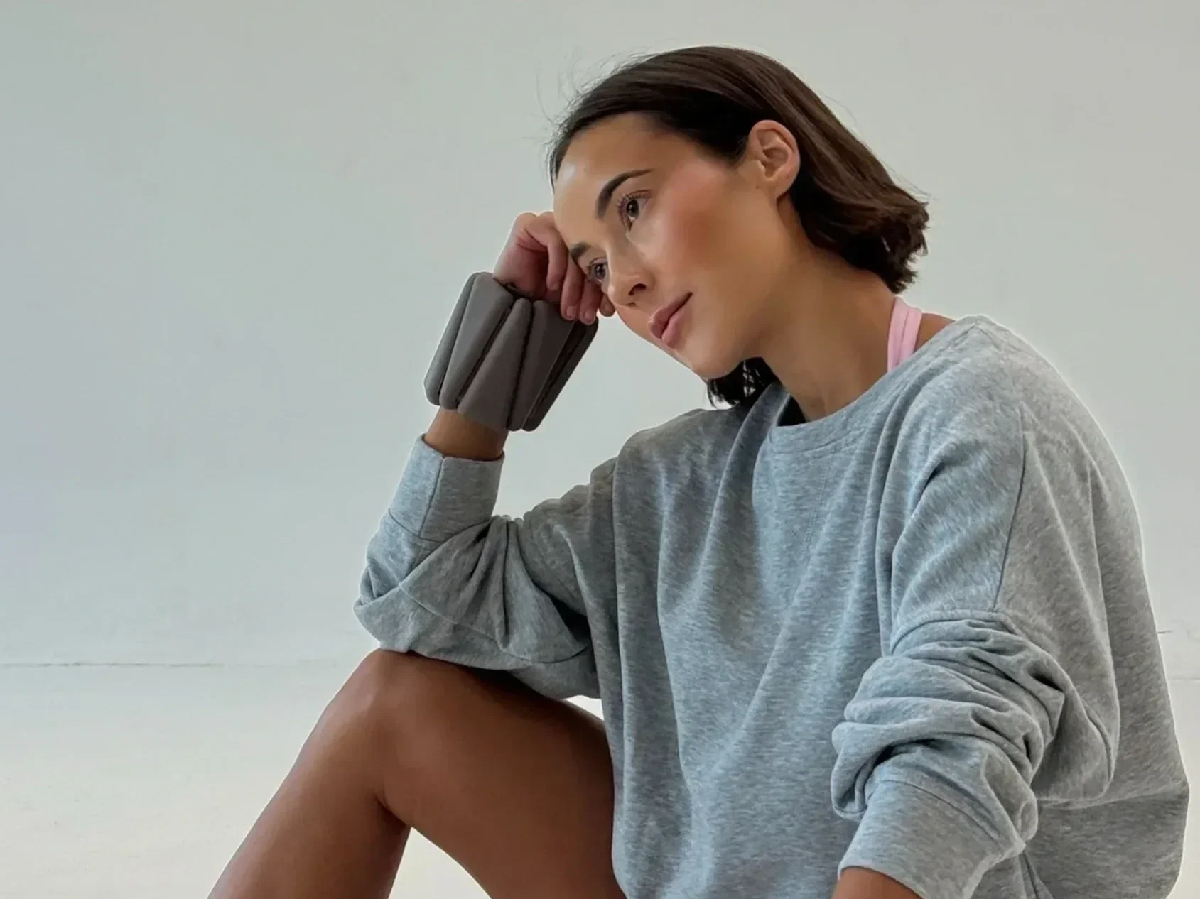 A young woman with shoulder-length brown hair sitting against a plain white wall, resting her head on her right hand with a large gray wrist brace, wearing a gray sweatshirt and pink sports bra, looking contemplative.