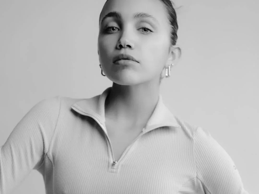 Black and white portrait of a young woman with her hair pulled back, wearing a zip-up top and earrings.