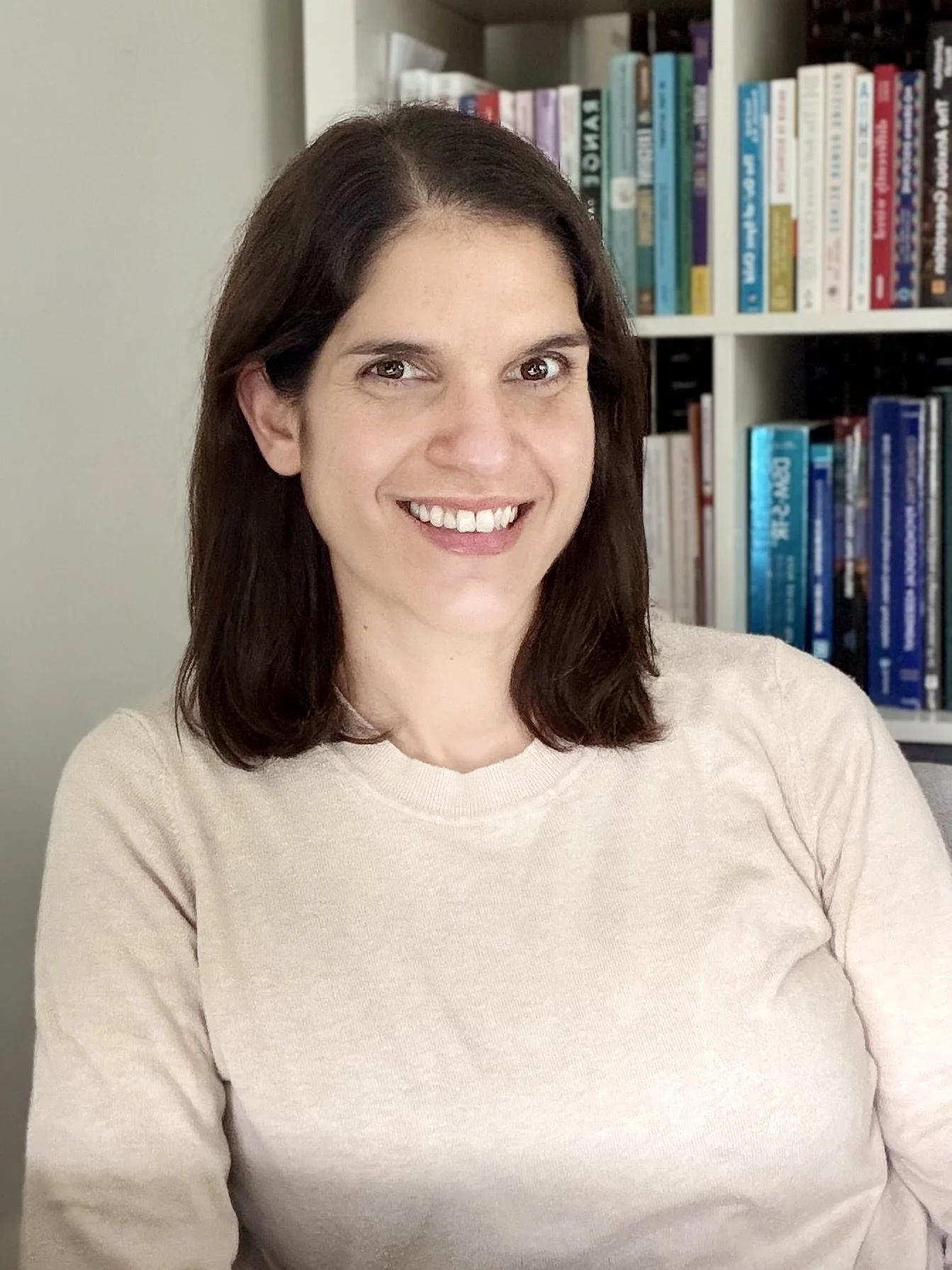 A smiling woman with dark brown hair and brown eyes, wearing a light beige sweater, standing in front of a bookshelf filled with books.