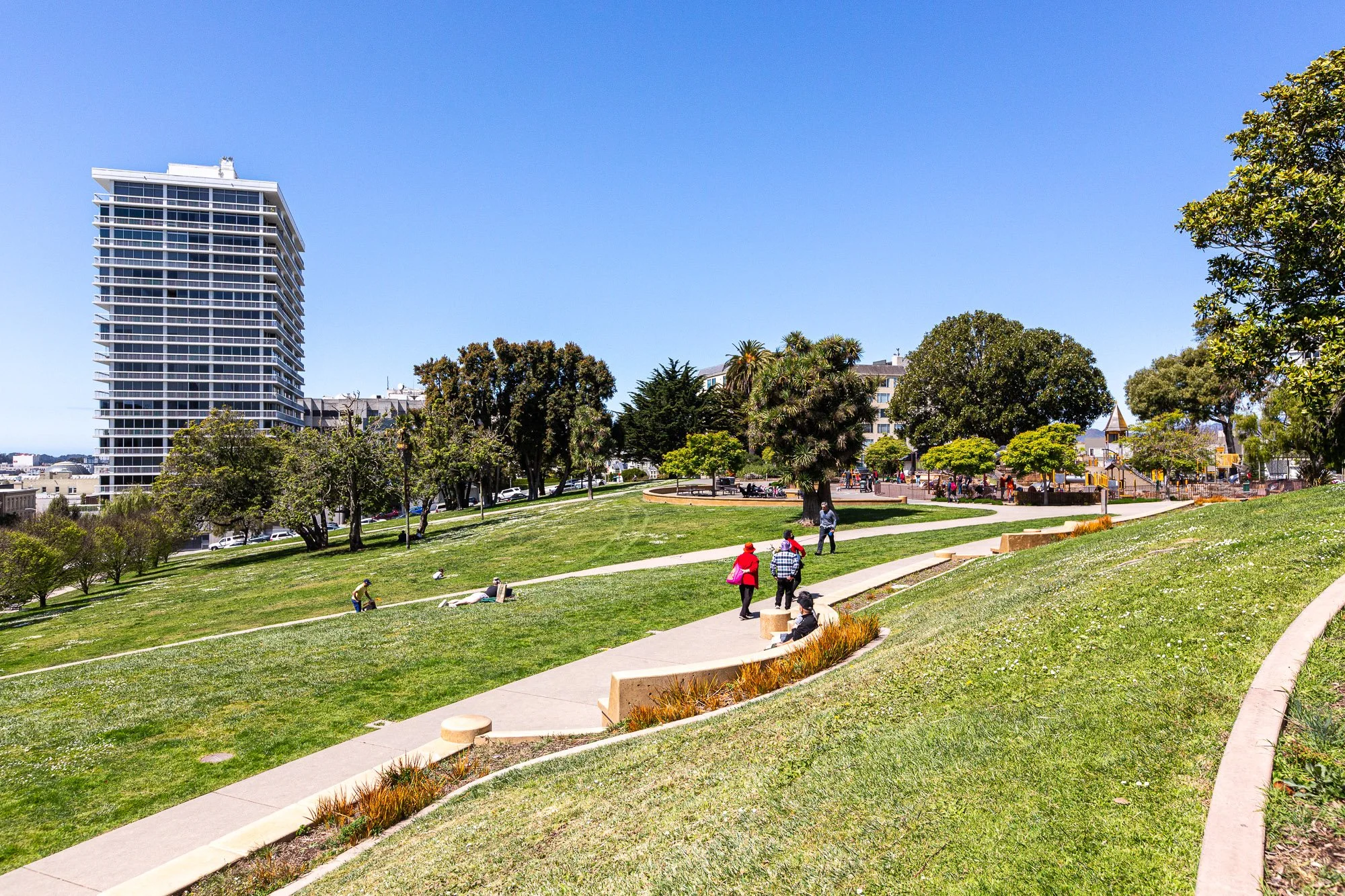 People walking and sitting on benches in a sunny park with green grass, trees, and a tall building in the background.