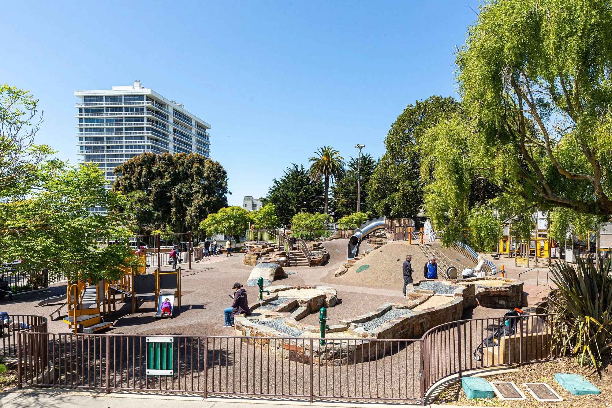 A city park with various playground equipment, trees, and a high-rise building in the background, under clear blue skies.