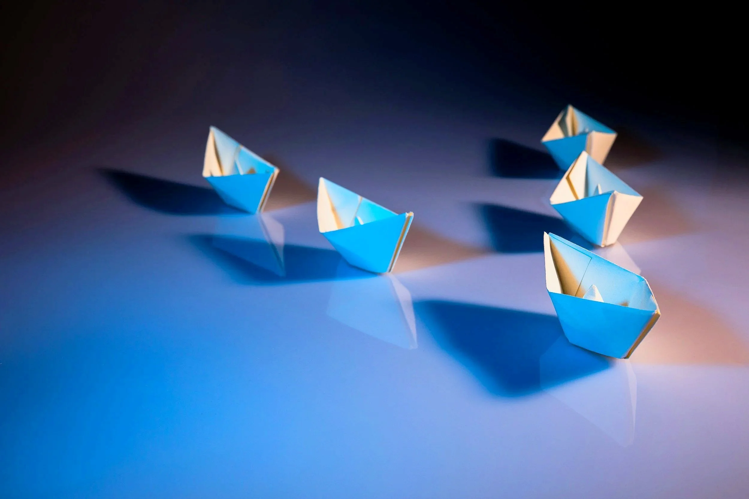 Six small paper boats, some blue and some white, casting shadows on a reflective surface with lighting creating a gradient background.