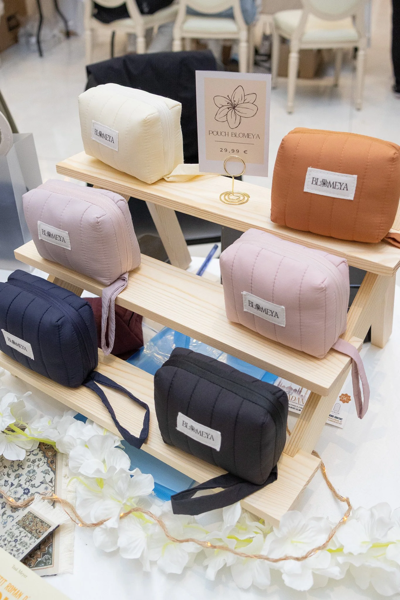 Différents pochons colorés en tissu, avec des étiquettes portant le nom 'BLOMEYA', exposés sur une étagère en bois à côté d'une carte de prix indiquant 'POCH BLOMEYA 29,99 €'.