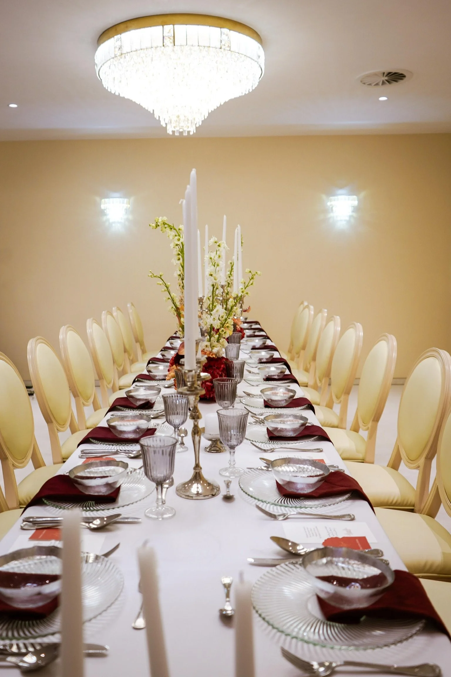 Salle à manger élégante avec un long table recouvert d'une nappe blanche, décorée de bougies, d'arbustes floraux et de vaisselle en argent avec des verres en cristal. Un lustre en cristal brillant est suspendu au plafond.
