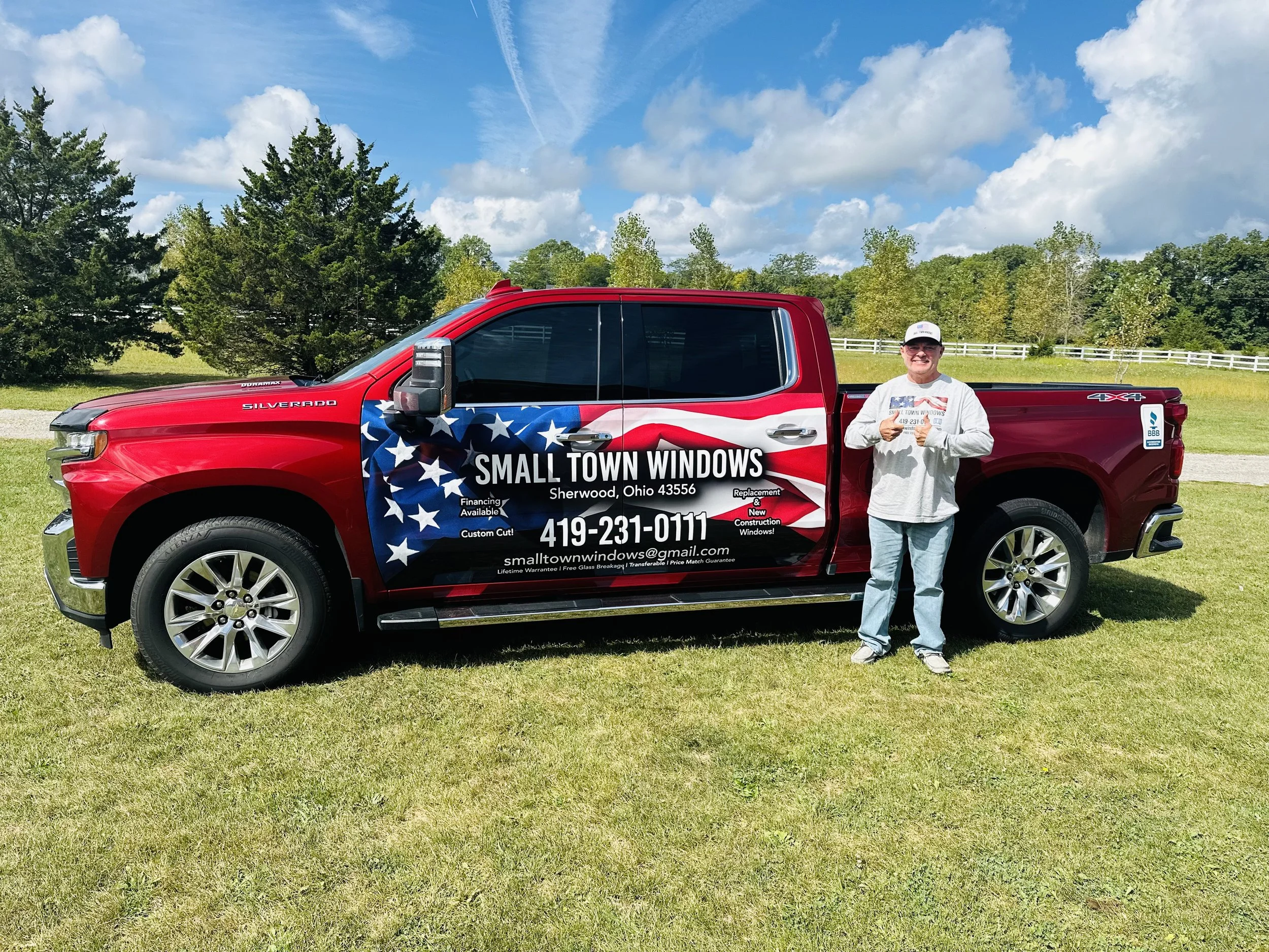 Man standing next to a red pickup truck with an American flag design advertising Small Town Windows, parked on grassy field with trees and blue sky in the background.