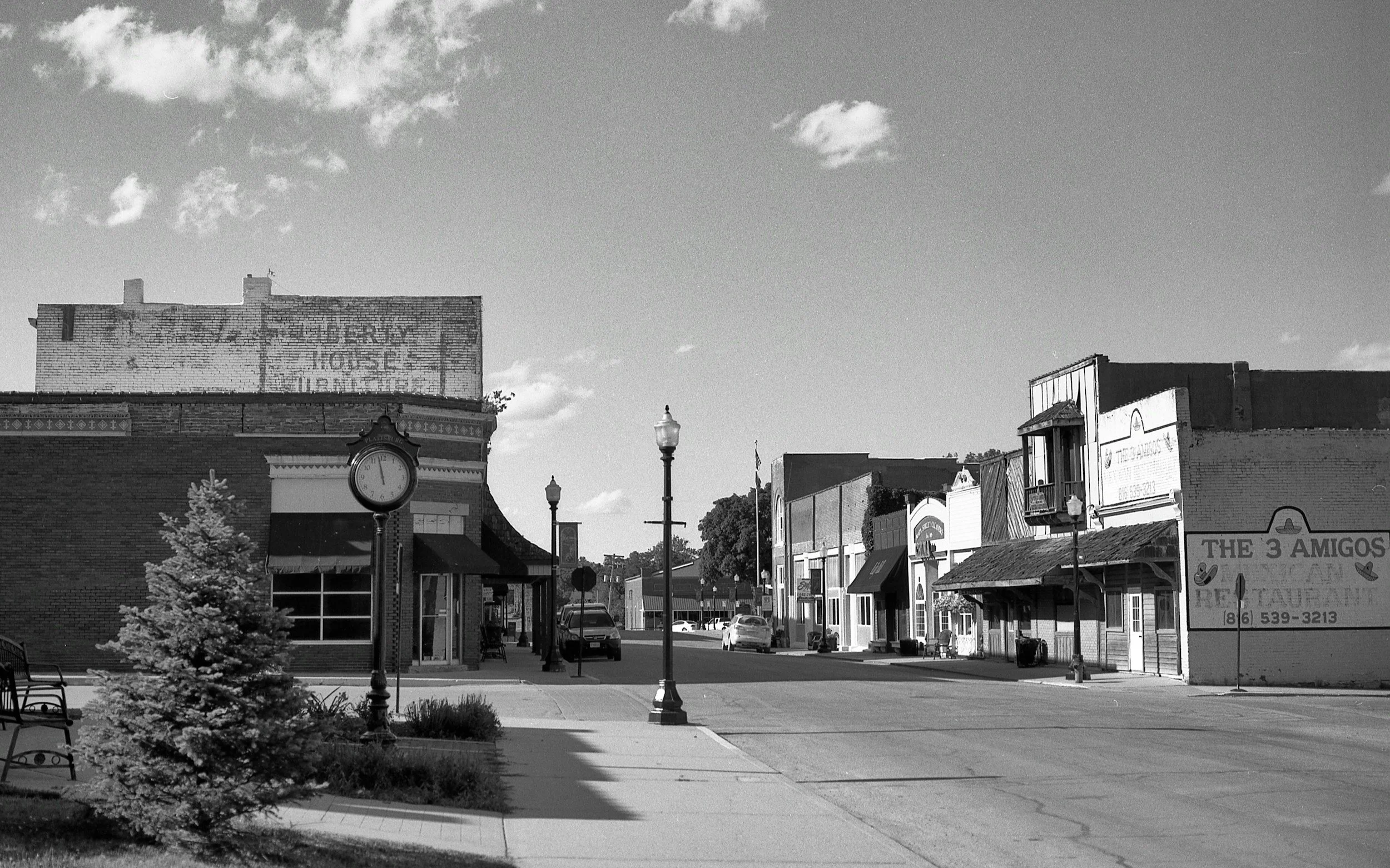 A black and white street scene with small shops, a street clock, street lamps, and parked cars. The sky has some clouds. Small town nostalgia