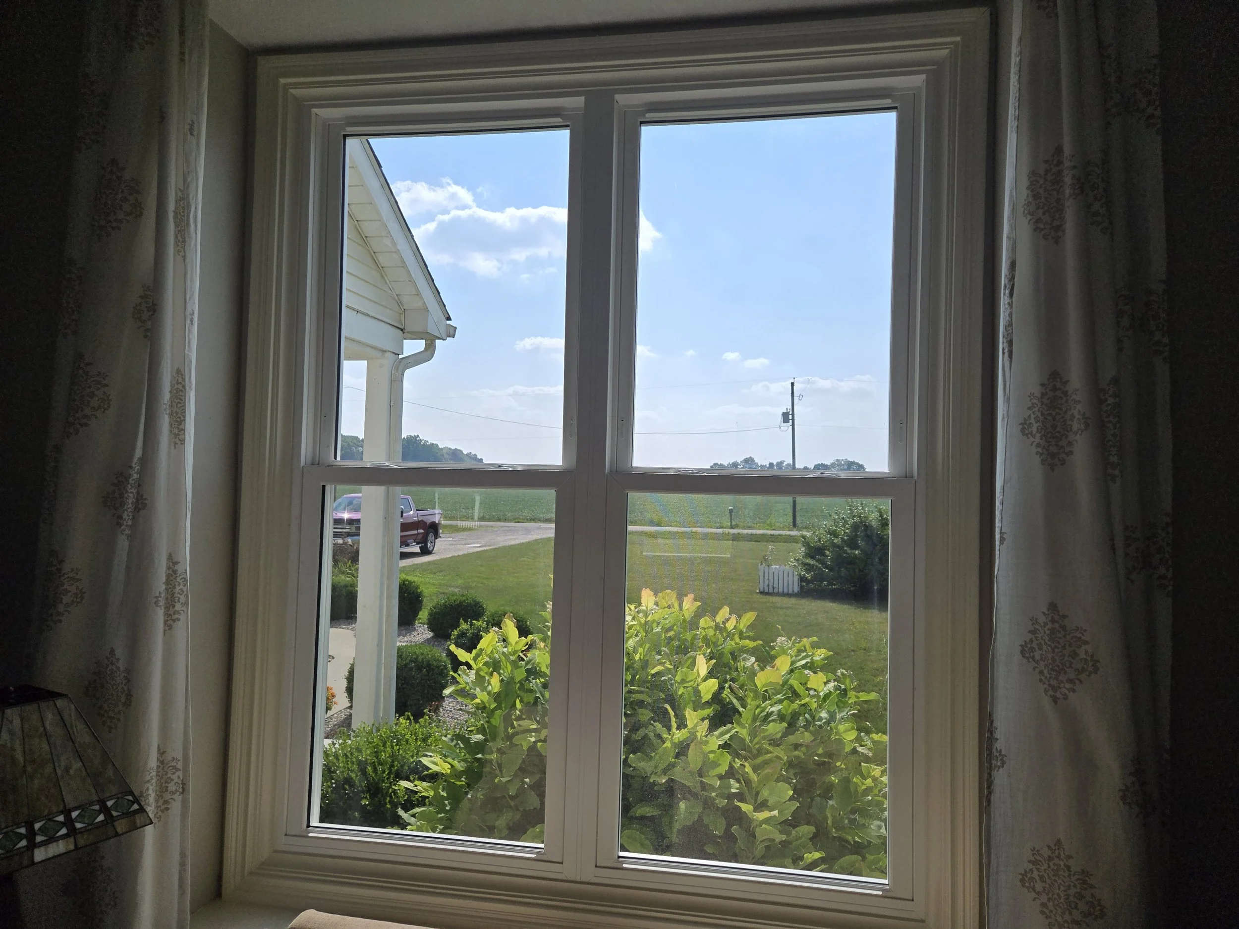 View through a window showing a driveway, a purple truck, green lawn, shrubs, a farm field, power lines, and a partly cloudy sky.