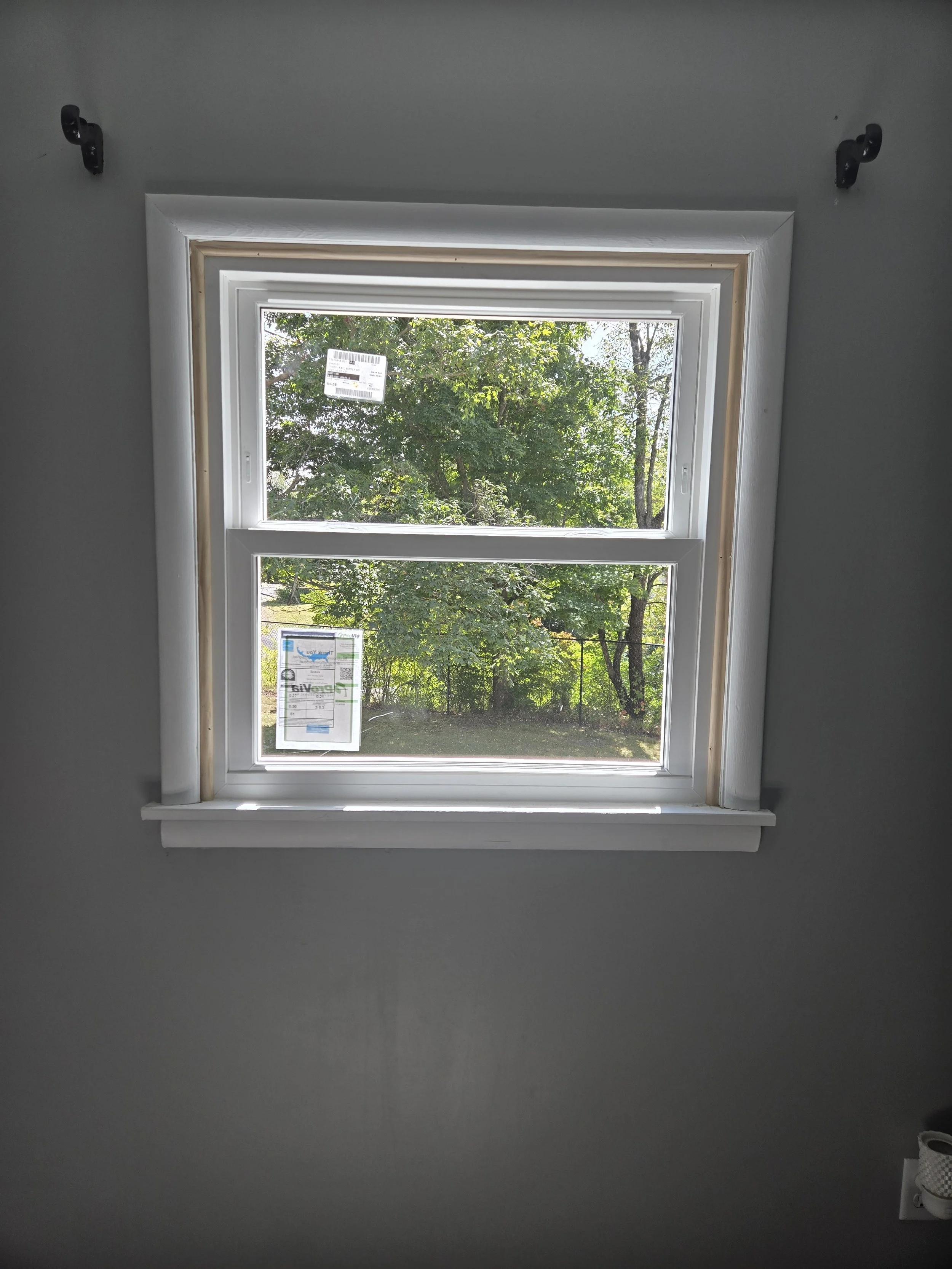 View of a double-hung window with white frame, showing trees and greenery outside, with a sticker on the upper pane and a label on the lower pane, inside a room with gray walls.