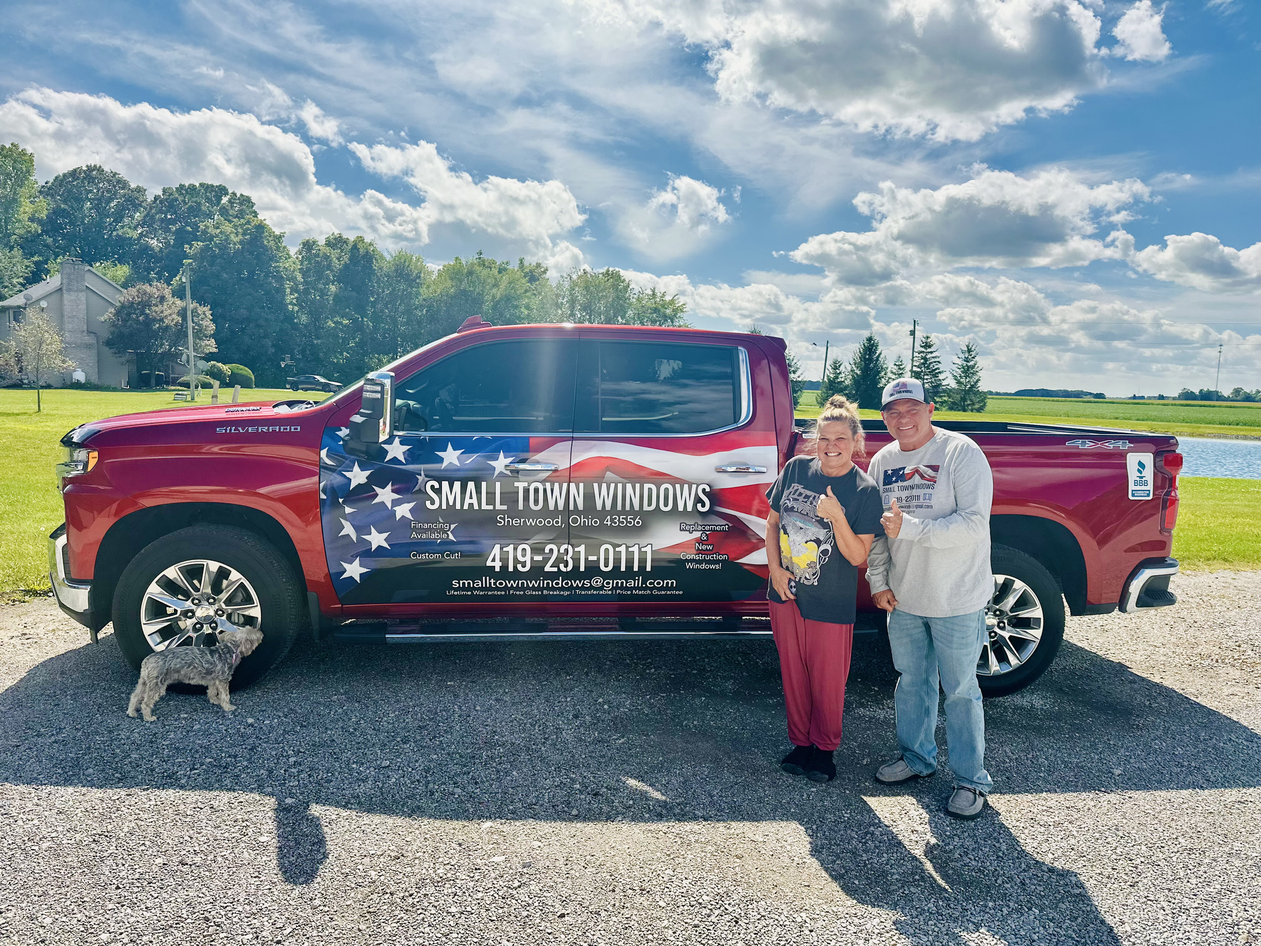 A man and woman standing next to a red truck with a patriotic design and advertising for Small Town Windows, with a small dog nearby on the ground, outdoors on a sunny day with a partly cloudy sky and green landscape in the background.