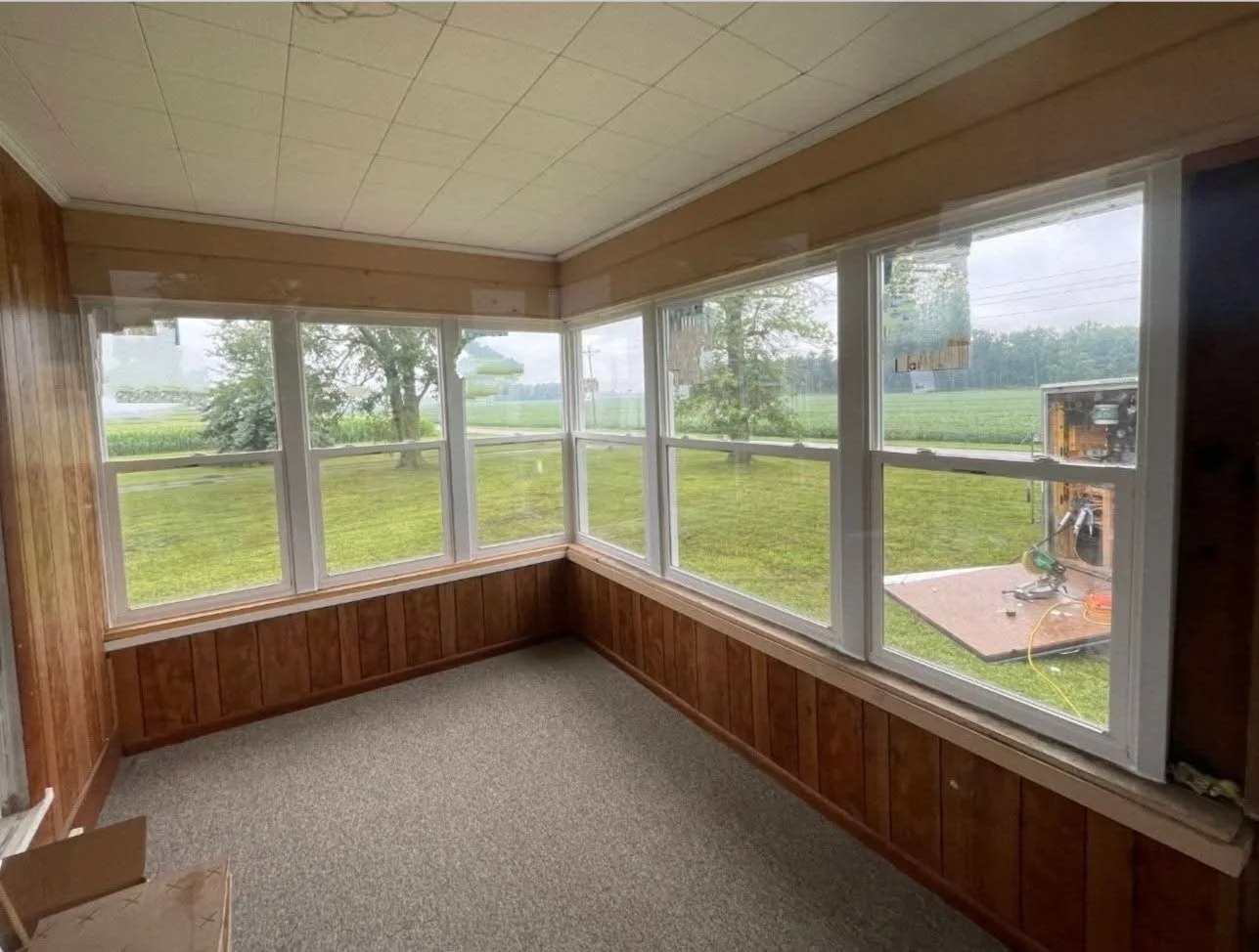 Sunroom with wood paneling and large windows showing green grass and trees outside.
