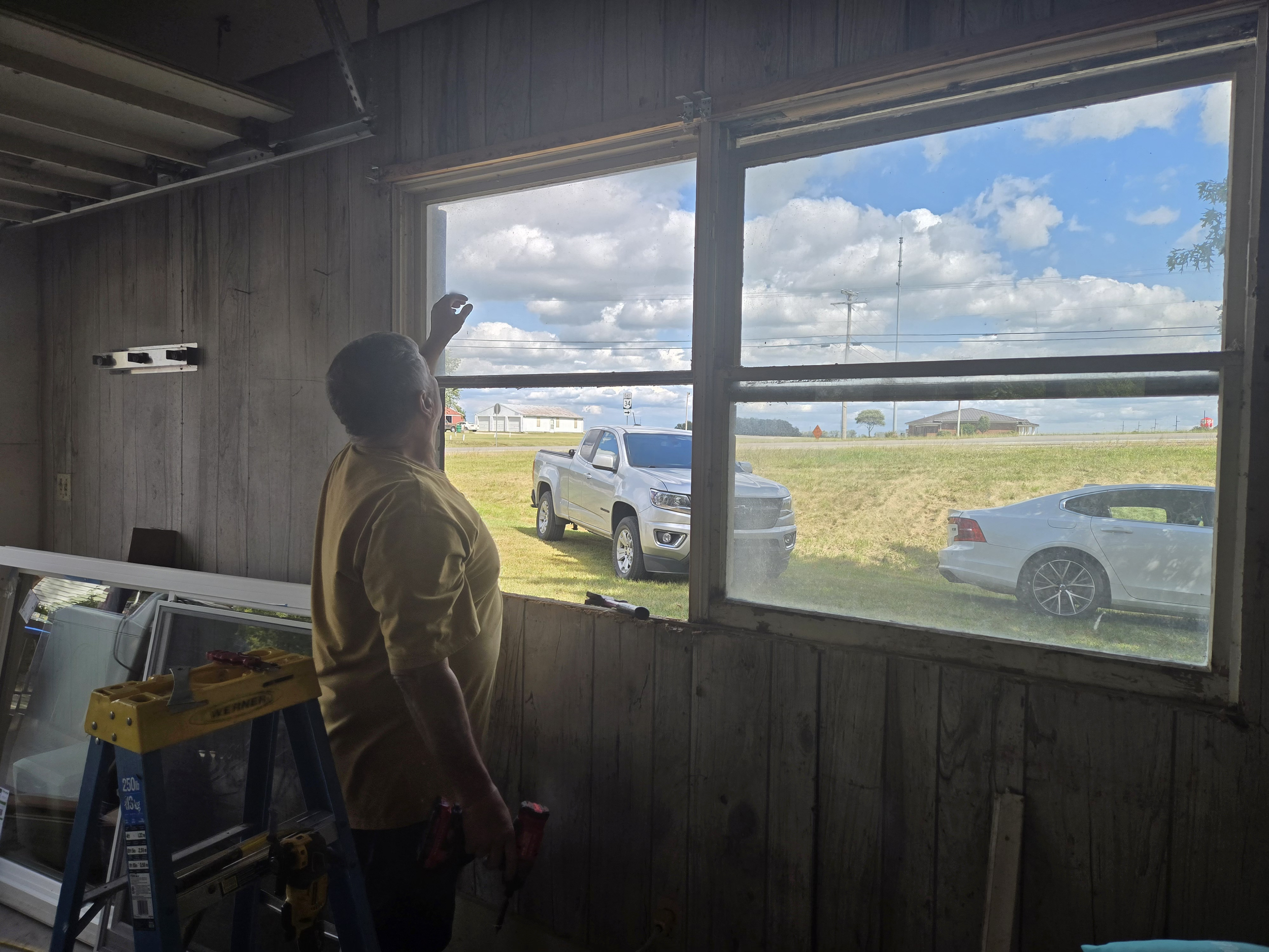 A man standing inside a room with wood-paneled walls, using a tool to work on the window frame. Outside, there are two parked cars on a grassy area, a pickup truck and a sedan, visible through the large window. The sky is partly cloudy with blue and
