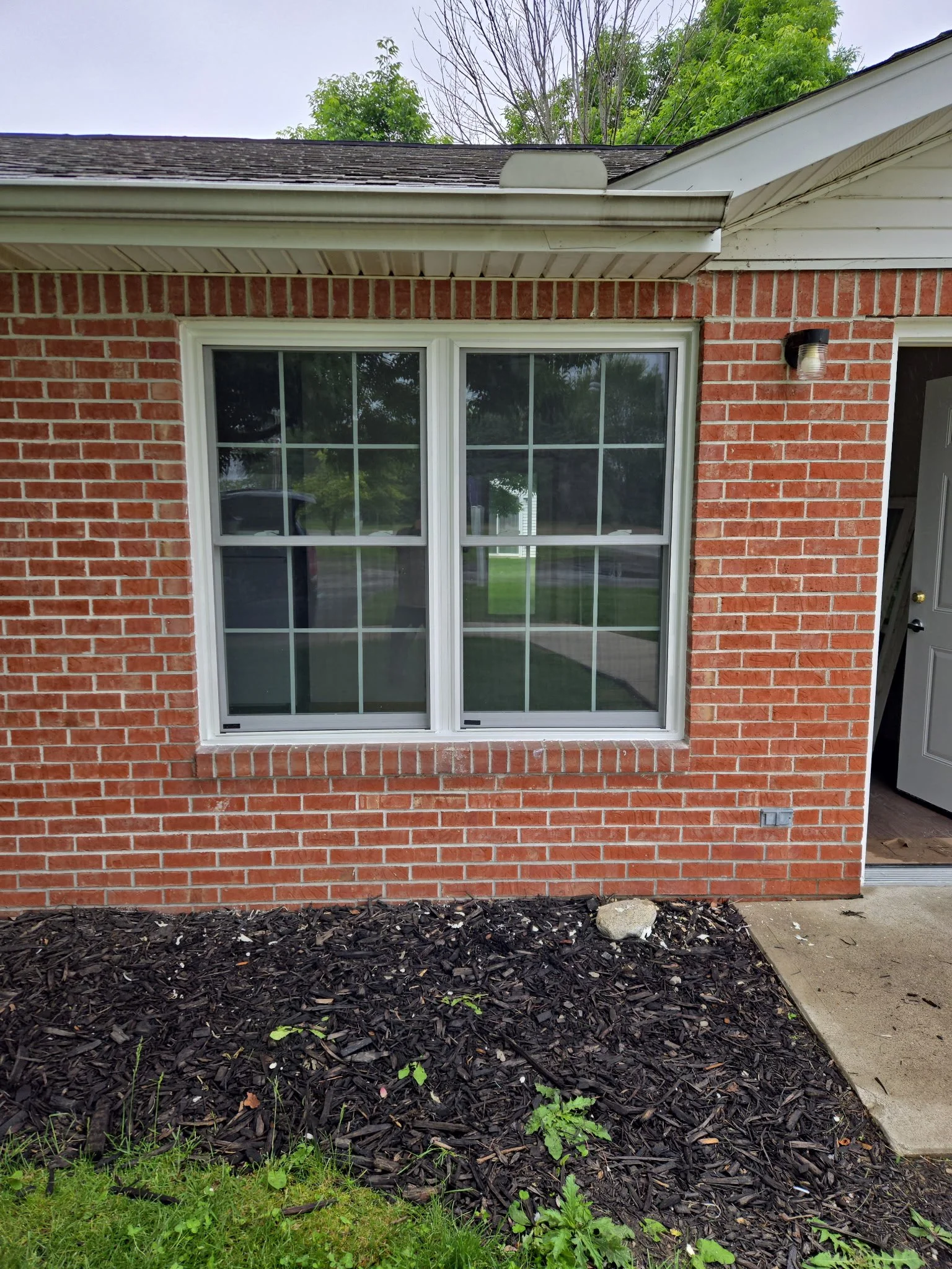 Front of a brick house with a large double window, a small outdoor light fixture, and a landscaped flower bed with black mulch and small plants.