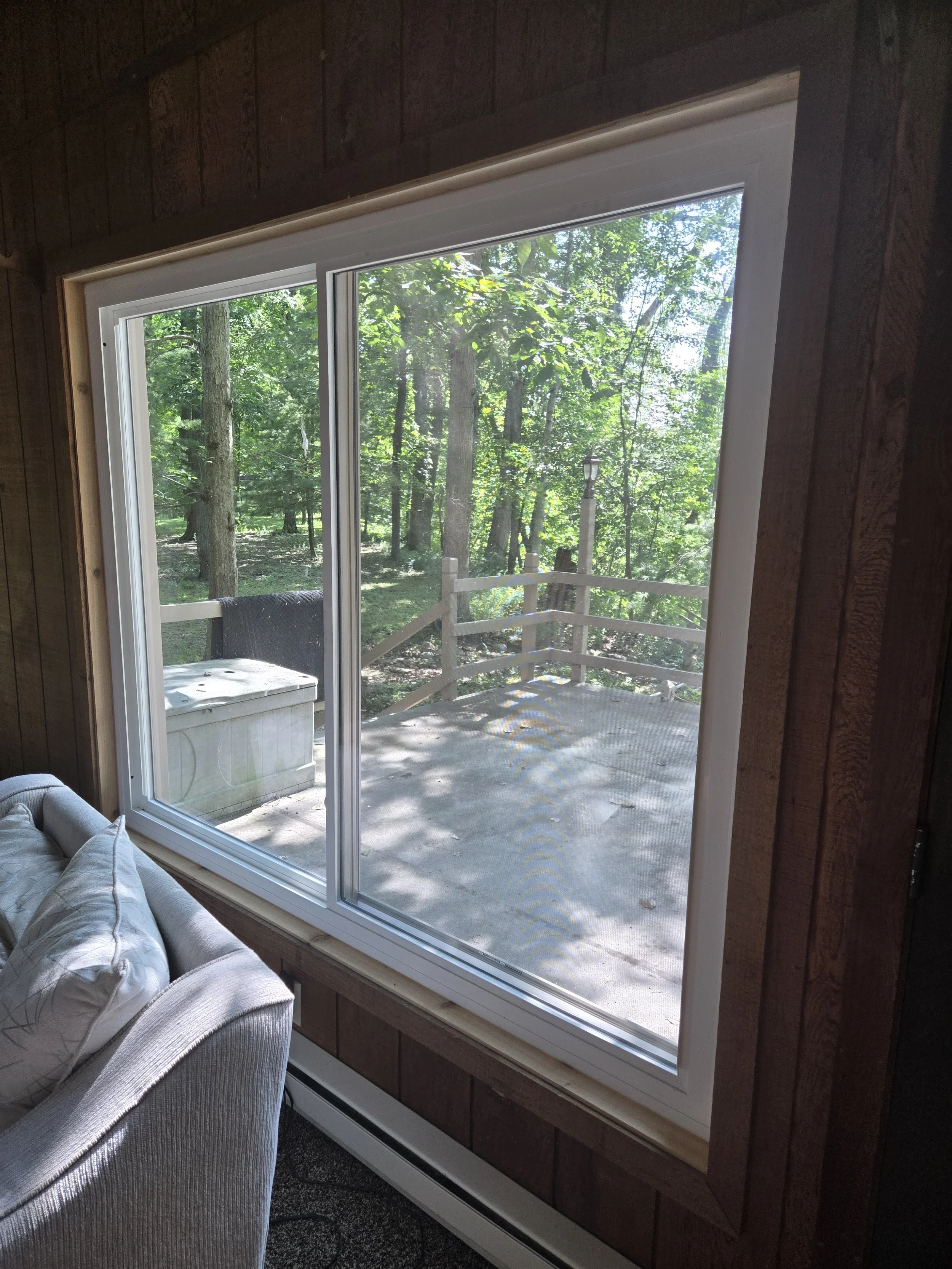 View of a wooden deck and lush green trees seen through a large window from inside a room with wooden walls and a beige sofa.