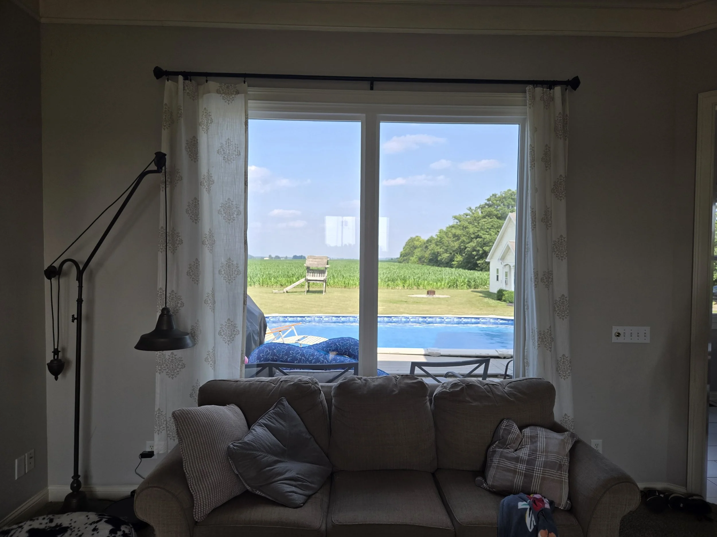 View through a living room window showing a backyard with a swimming pool, green field, and blue sky with clouds.