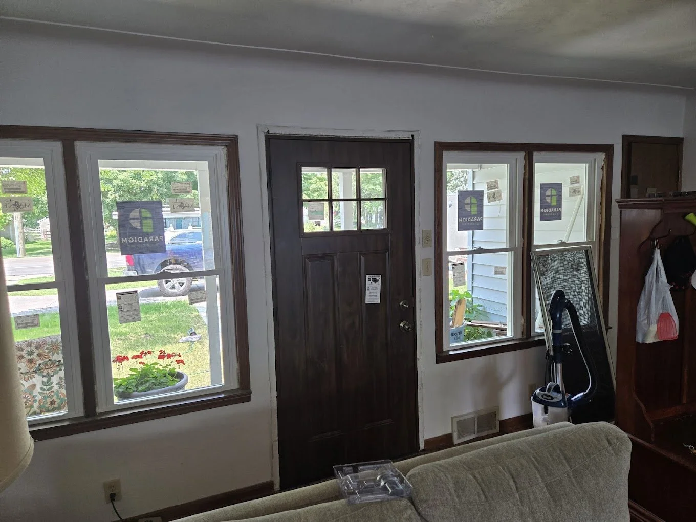 Interior view of a house entrance with three windows, a dark wooden front door with a small window, and a glimpse of the outside yard and street. There is a couch partially visible at the bottom of the image, and some household items including a vacu