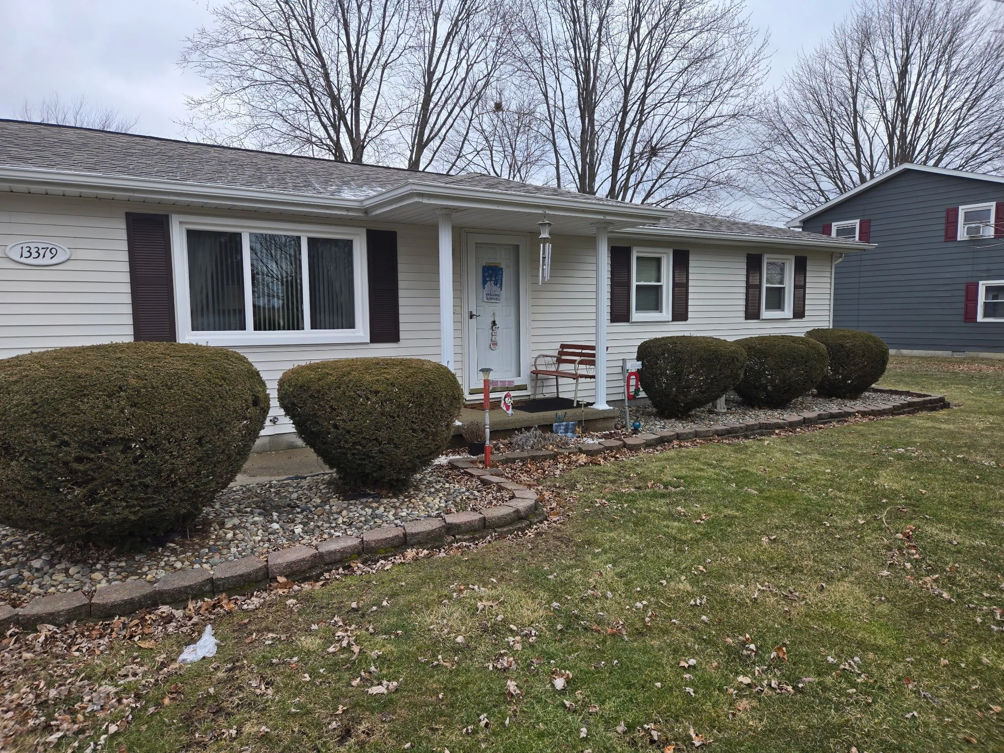 Front yard of a house with white siding, black shutters, trimmed bushes, and a small porch with a wooden bench. The house number is 13379, and there are leafless trees and another house in the background.