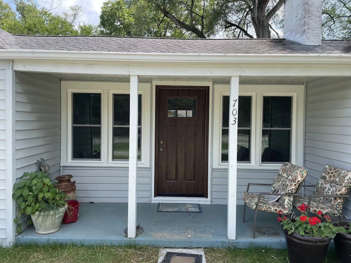 Front porch of a house with a wooden door, two large windows, potted plants, and patio chairs on a concrete porch. House number '703' is visible on a white support post.
