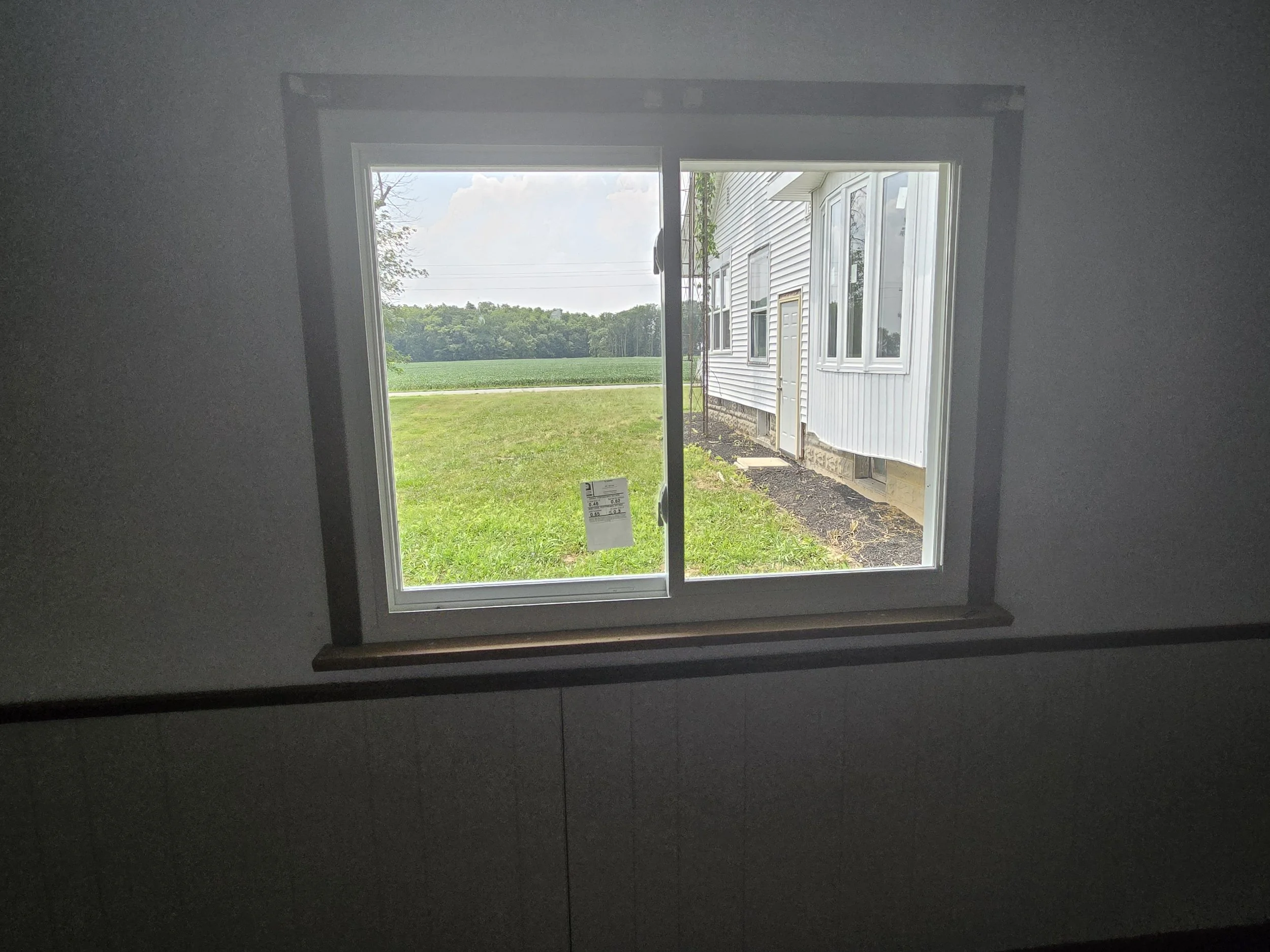 View through a window showing a grassy lawn, a tree line, and a cloudy sky outside a white house with multiple windows.