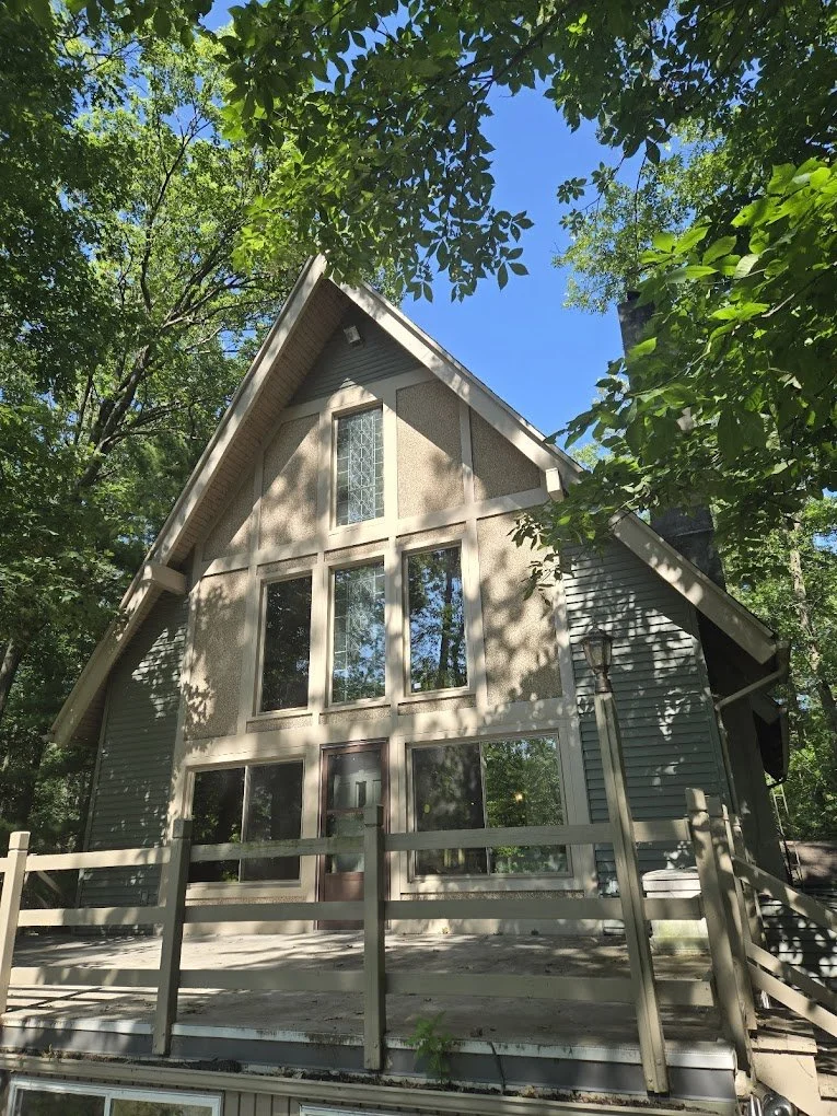 A two-story house with a steep gable roof and large front windows, surrounded by tall green trees and a wooden deck in a forested area under a clear blue sky.