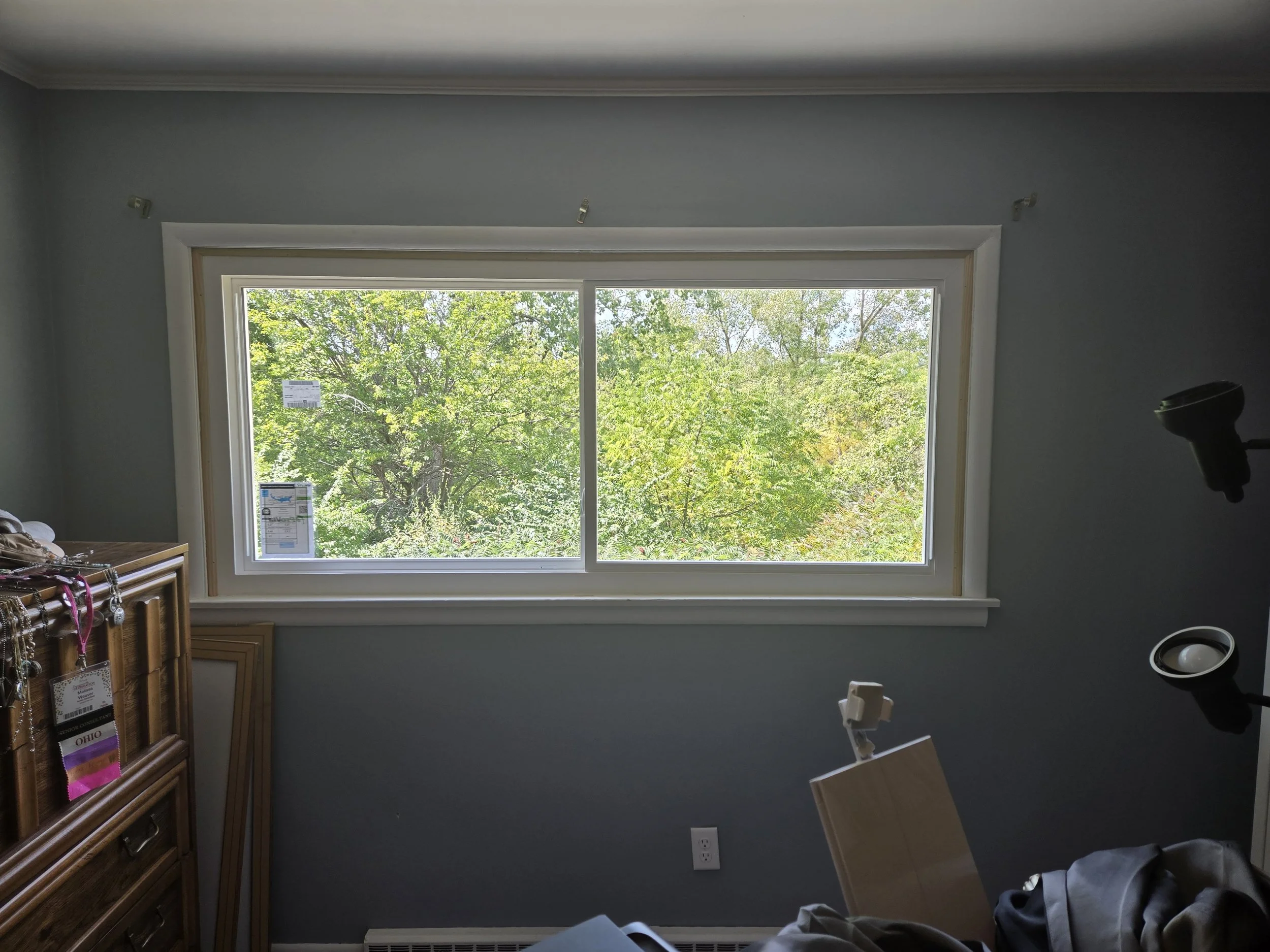 Empty room with a large window showing green trees outside, a wooden dresser, and various items on the floor and furniture.